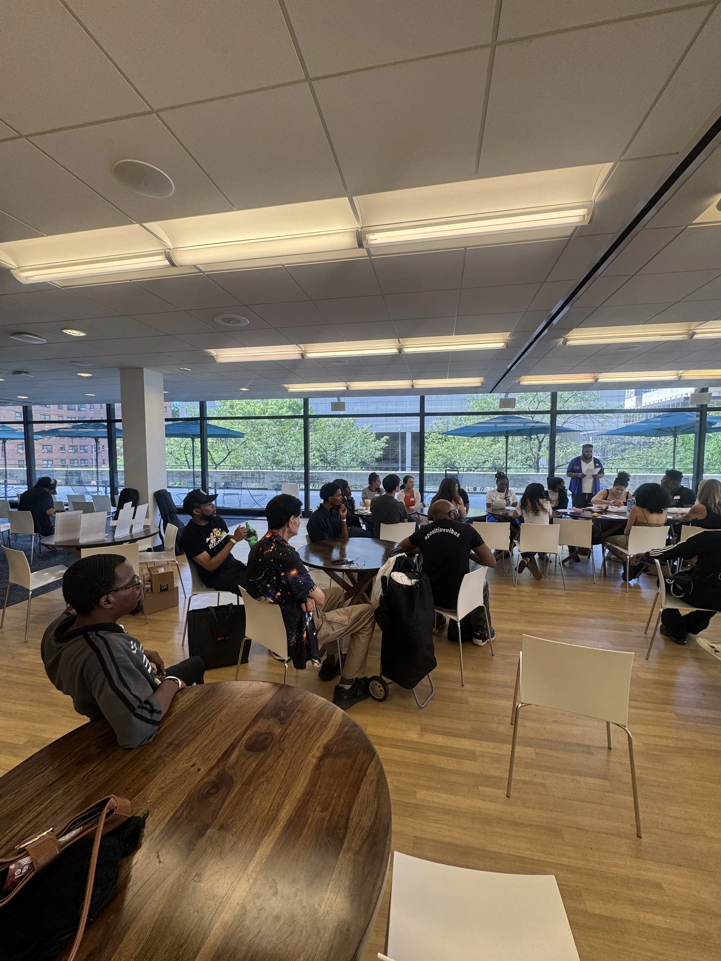People sitting and standing in a spacious room with large windows, fluorescent lights on the ceiling, and green trees outside.