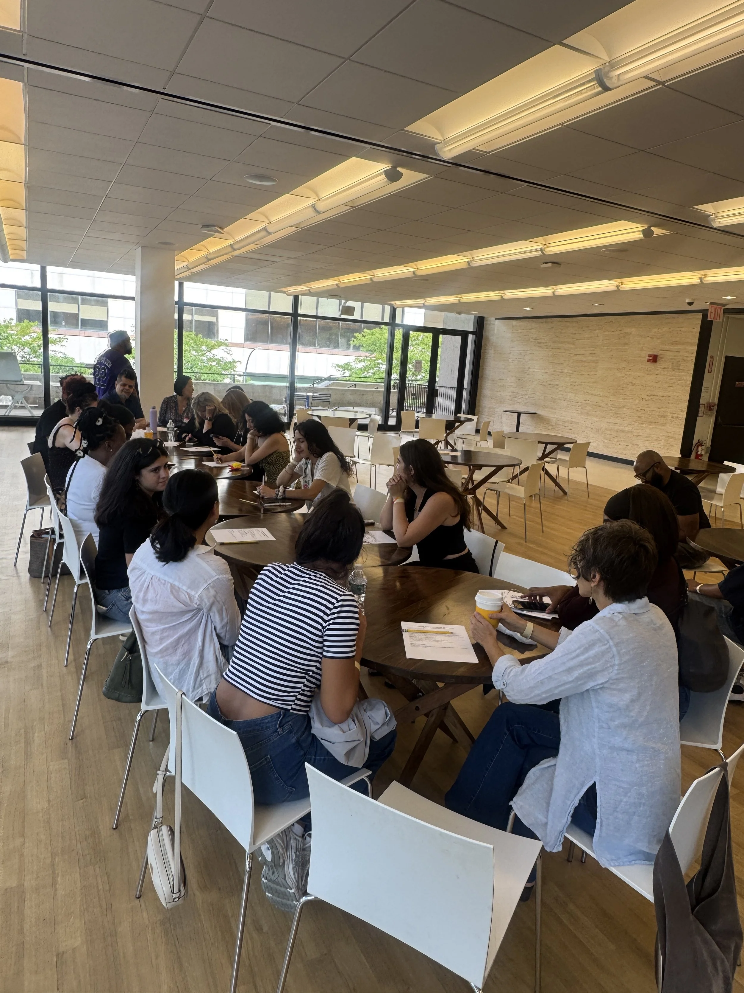 Group of people sitting around a long table in a bright room with large windows, some looking at their phones and papers, and one person holding a cup.