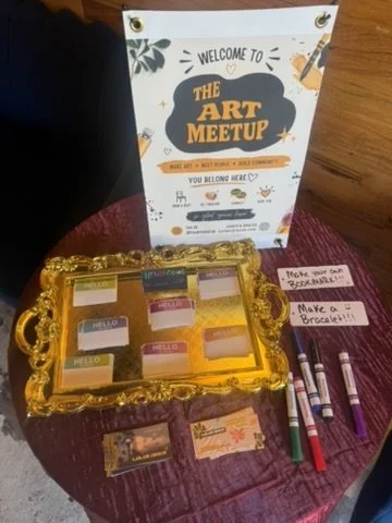 Display table with a sign saying 'Welcome to the Art Meetup,' a gold decorative tray with small jars of paint or ink, pens, and notes encouraging making bracelets or brooches.