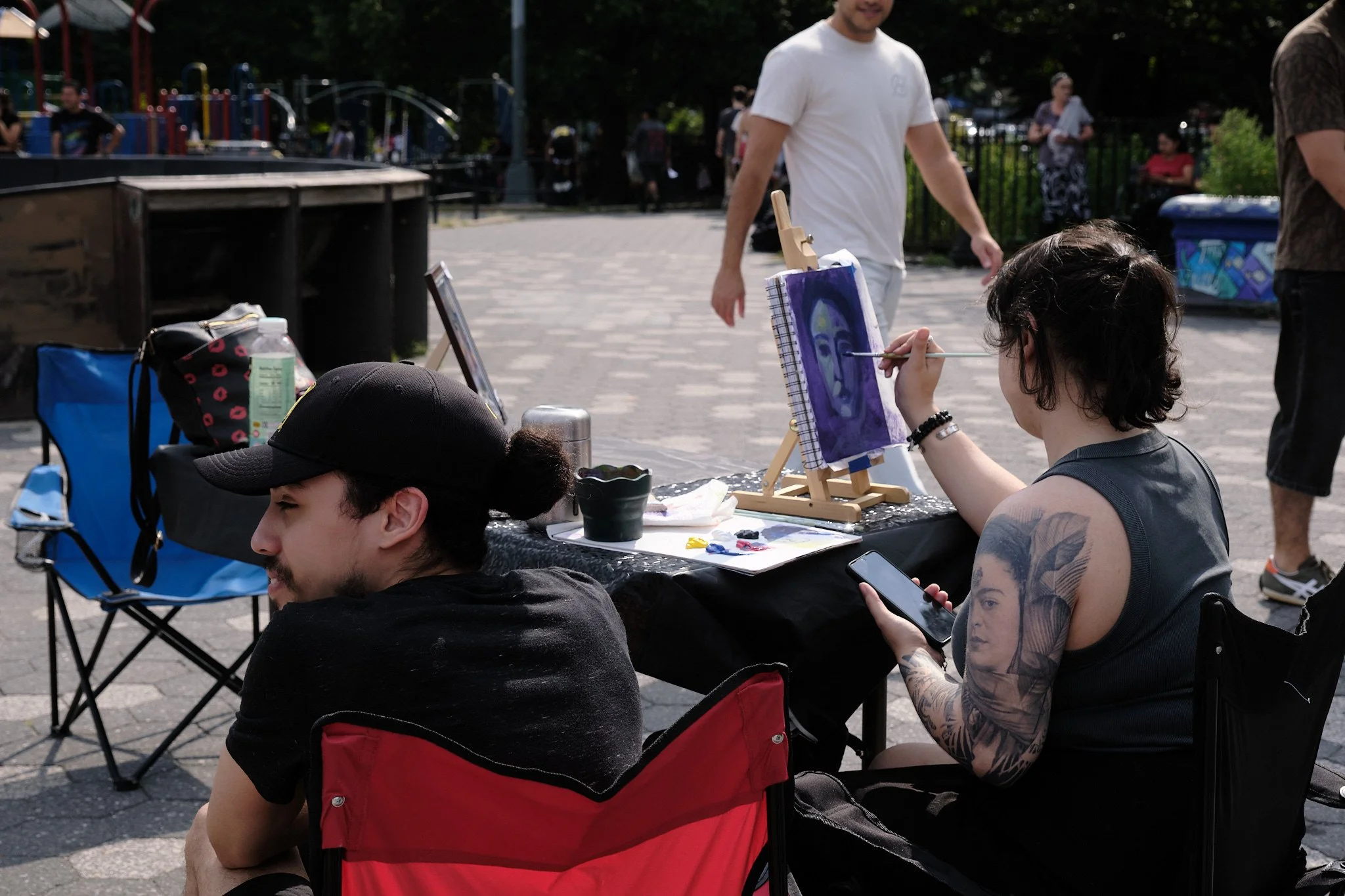 A woman with tattoos on her arm painting a portrait on a small easel at an outdoor event. She is seated at a black table with various art supplies and mobile device nearby. A man with a beard in a white t-shirt stands in the background with other peo