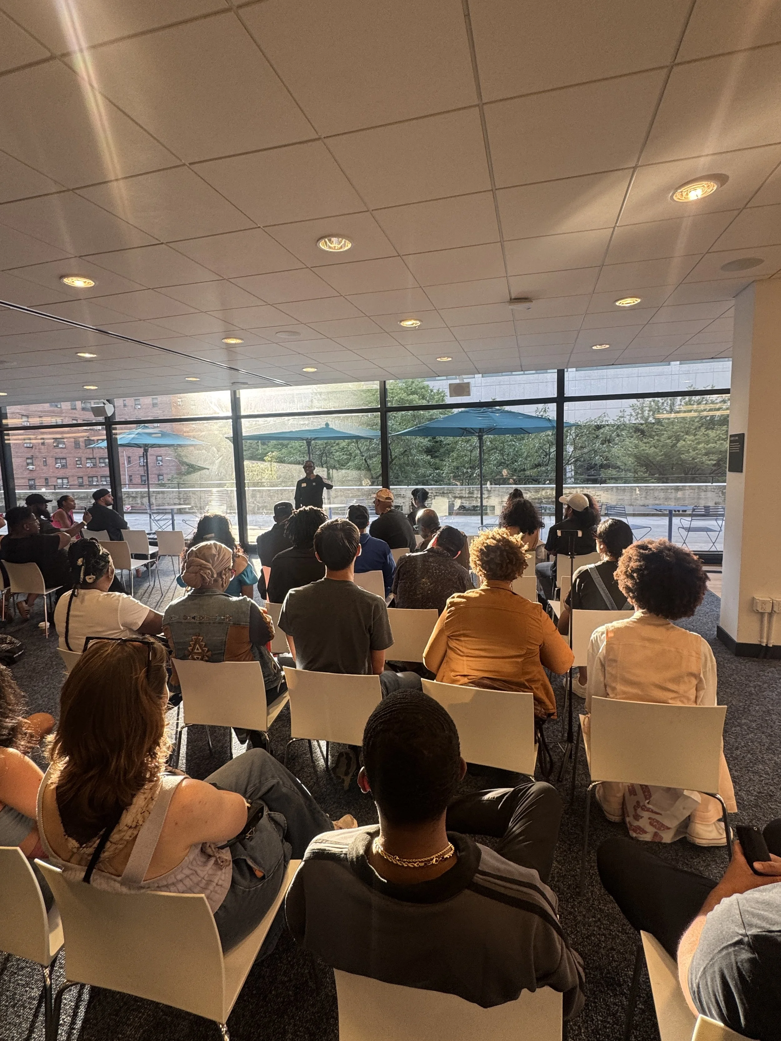 A group of people seated in chairs inside a room with large floor-to-ceiling windows, watching a presentation given by a woman standing near the windows. The room is well-lit with sunlight and has a modern design with white ceiling tiles and blue umb