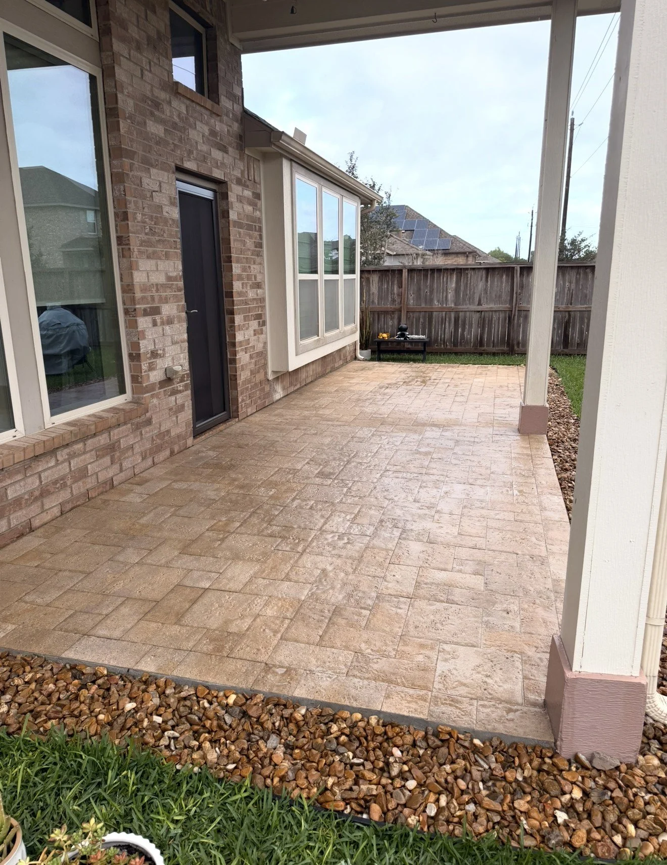 Clean brick patio with beige pavers, edged with small rocks and a patch of grass, part of a backyard with a wooden fence, neighboring houses, and solar panels on rooftops.