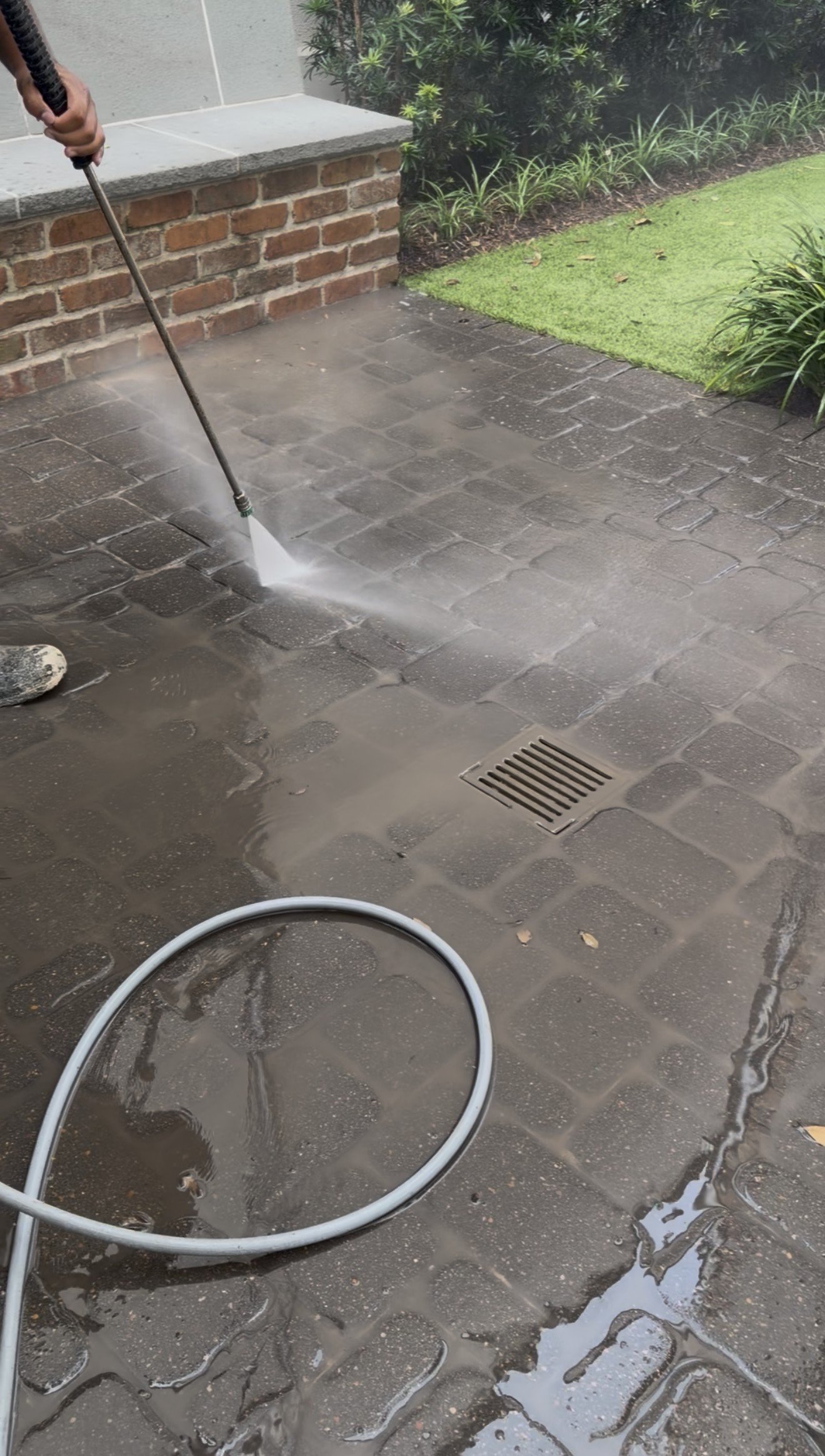 A person is pressure washing a brick patio outside near a garden bed with plants and grass. The water and soap from the cleaning process are visible on the dark stone tiling.