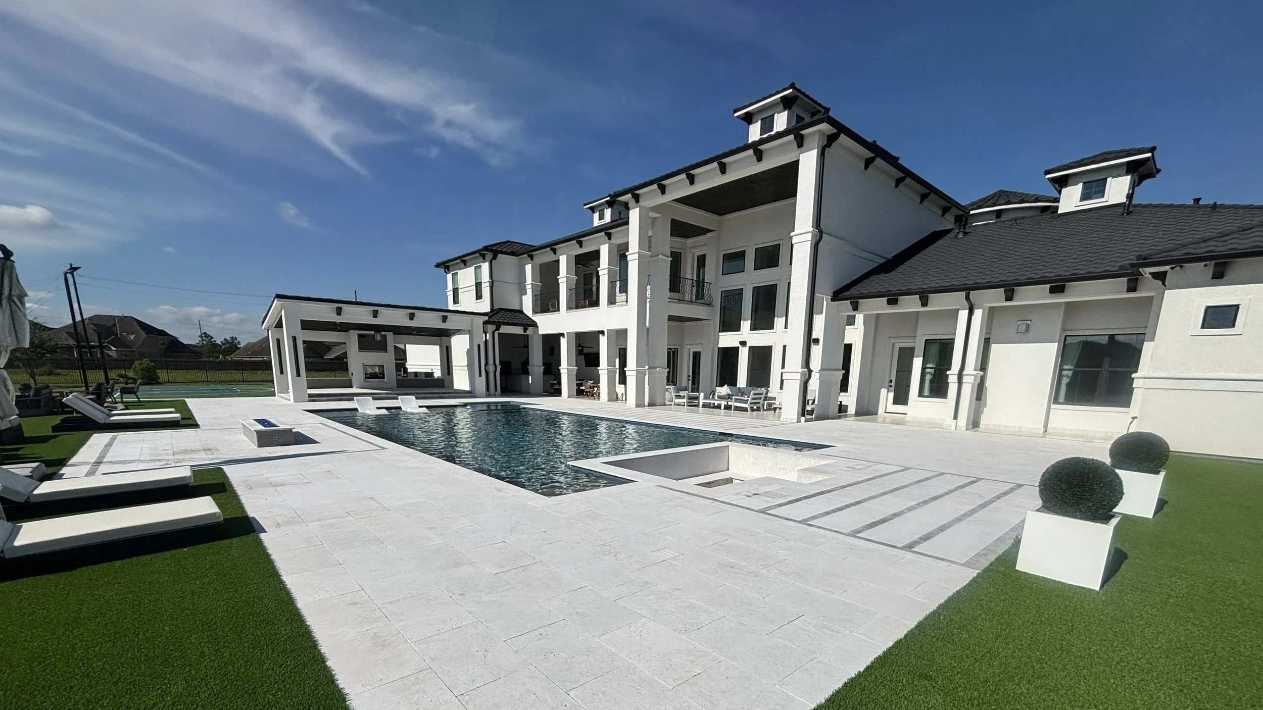 A luxurious backyard with a pool, lounge chairs, and a large white house with multiple levels and dark roofing. The area is well-maintained with artificial grass and modern decor, under a blue sky with few clouds.