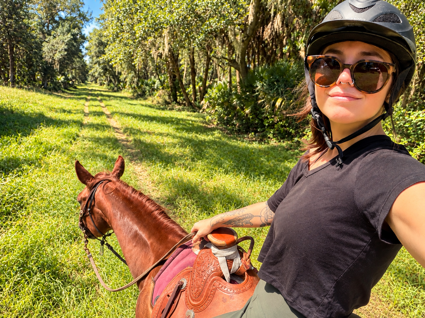 Woman wearing a bicycle helmet and sunglasses riding a horse on a grassy trail in a lush, green, wooded area.