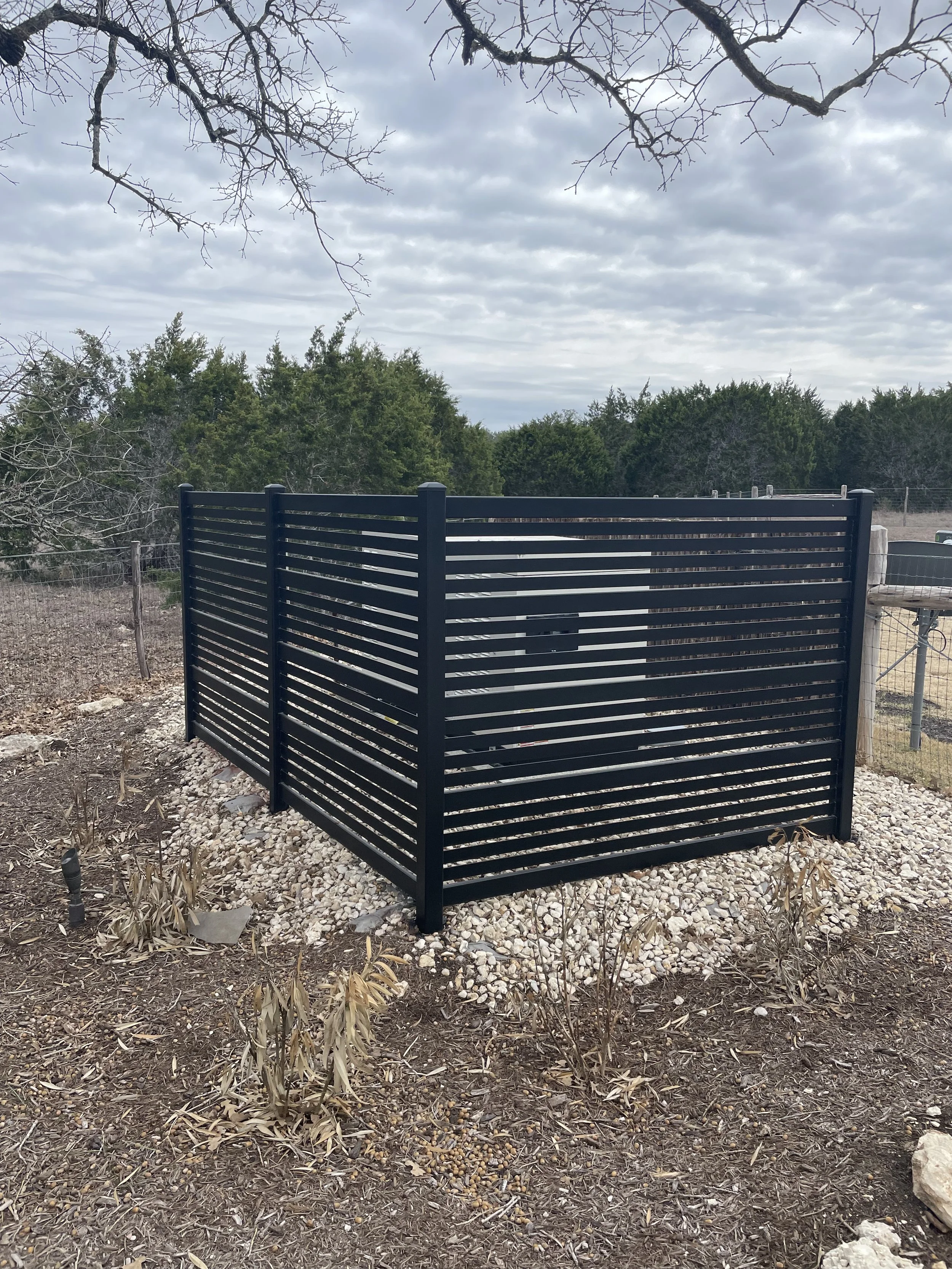 A black metal fence enclosure with horizontal slats screening a back up generator for a home.