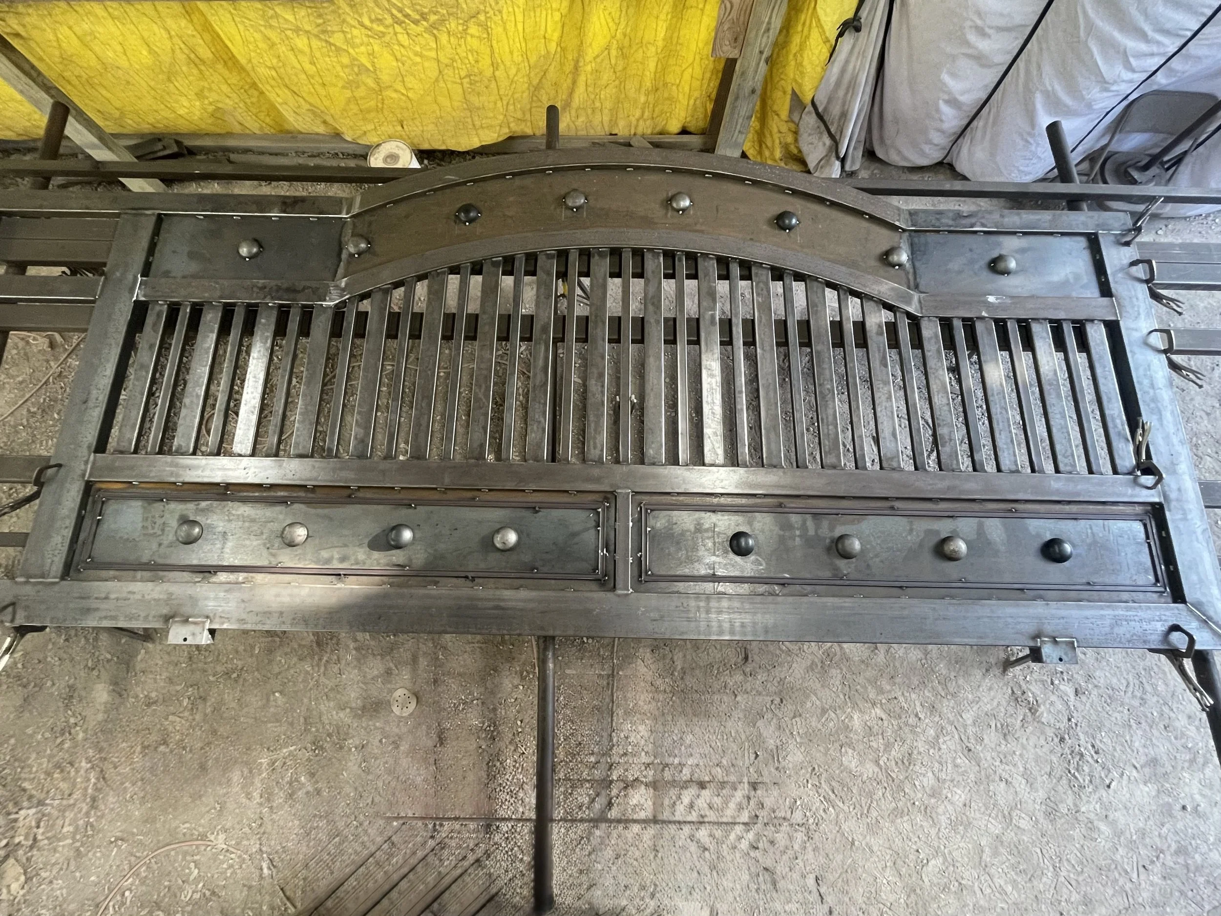 A metal decorative piece with a curved top, rivets, and vertical bars, situated on a dirt ground with some tools and fabric coverings in the background.