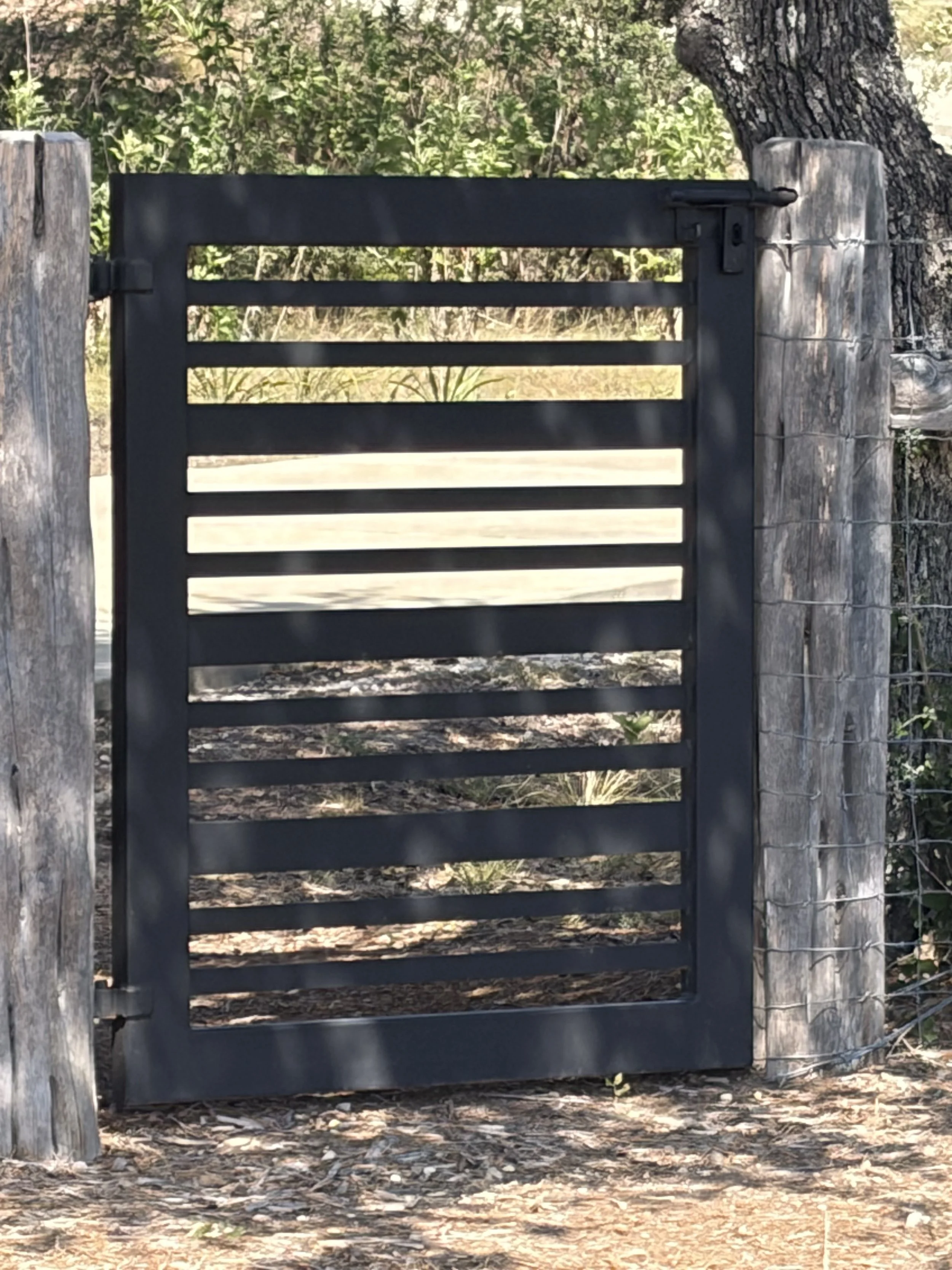 A black metal gate with horizontal slats, mounted between weathered wooden posts, in a natural outdoor setting with trees and bushes in the background.