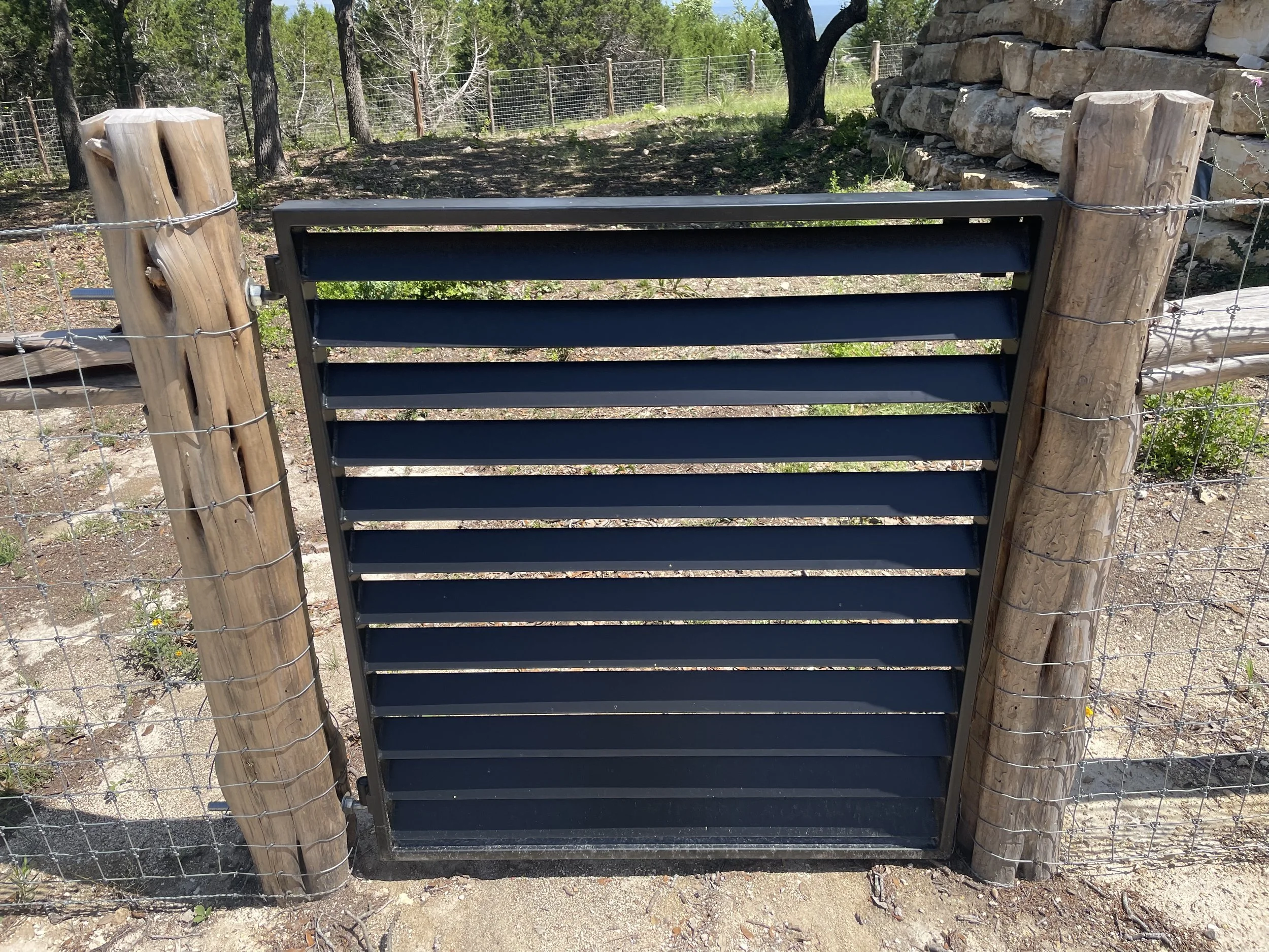 A metal gate attached between two wooden posts with barbed wire, set in a dirt area with trees and a stone wall in the background.