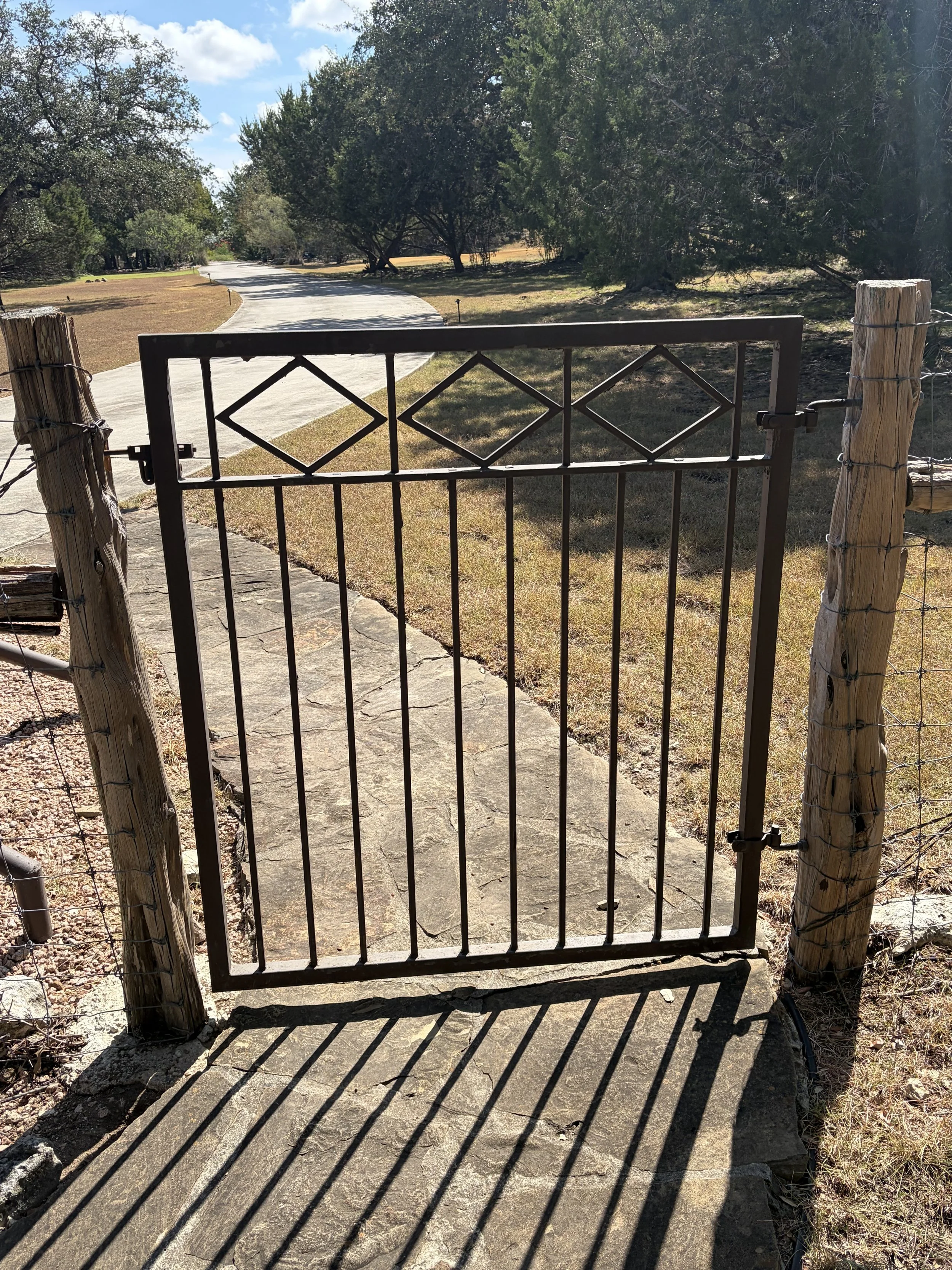 A black metal gate with diamond-shaped patterns, attached to wooden fence posts with barbed wire, opens onto a concrete pathway in a park-like setting with trees and grass.