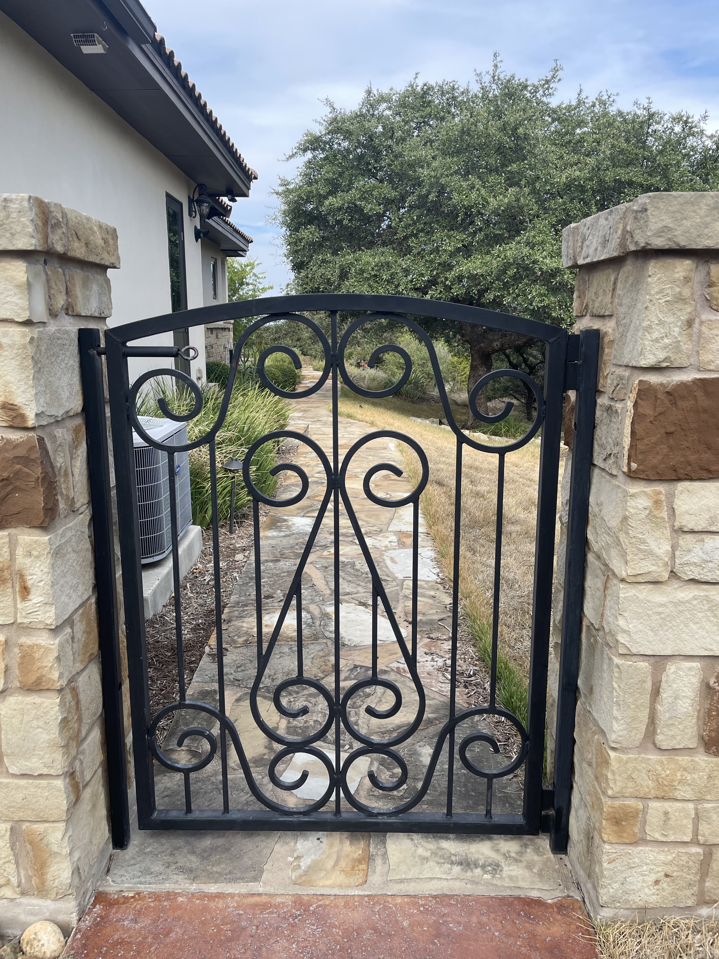 A decorative black metal gate with swirling design, flanked by stone pillars and leading to a stone pathway beside a white house with a tiled roof and an overgrown garden.