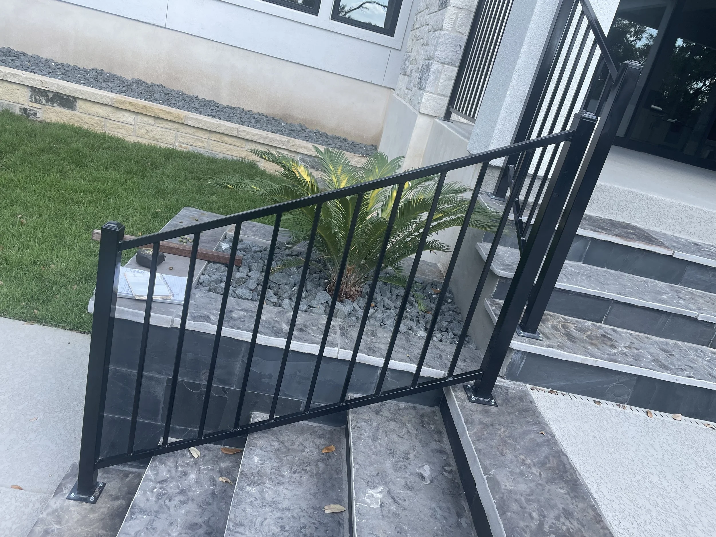 Front porch with black metal railing and steps, small shrubbery, and gray stone landscaping next to the house wall.