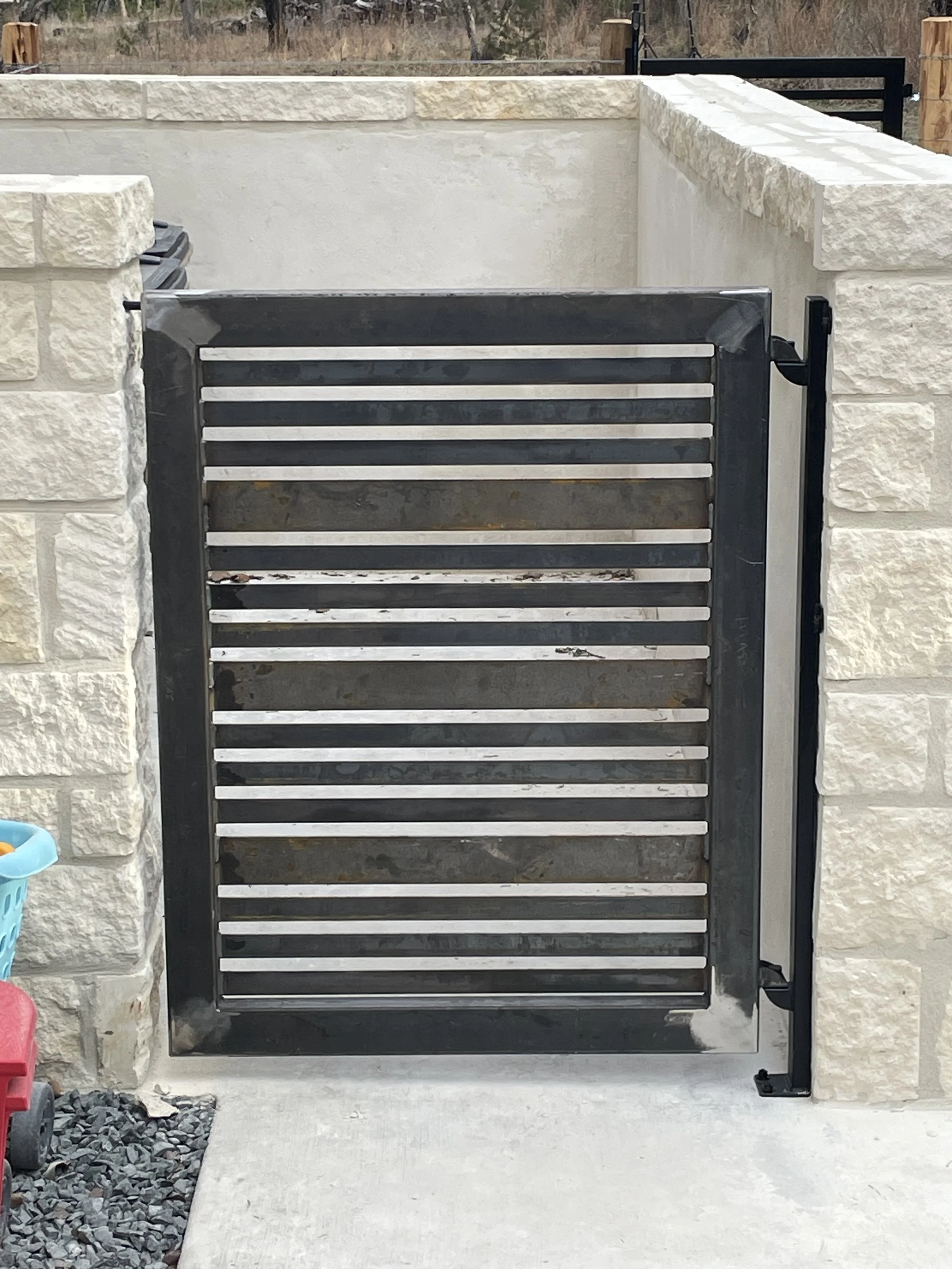 Image shows a metal horizontal slat gate in front of a beige stone wall that encloses and screens trash receptcales with a concrete pathway leading up to it.   is mounted on a black metal post.