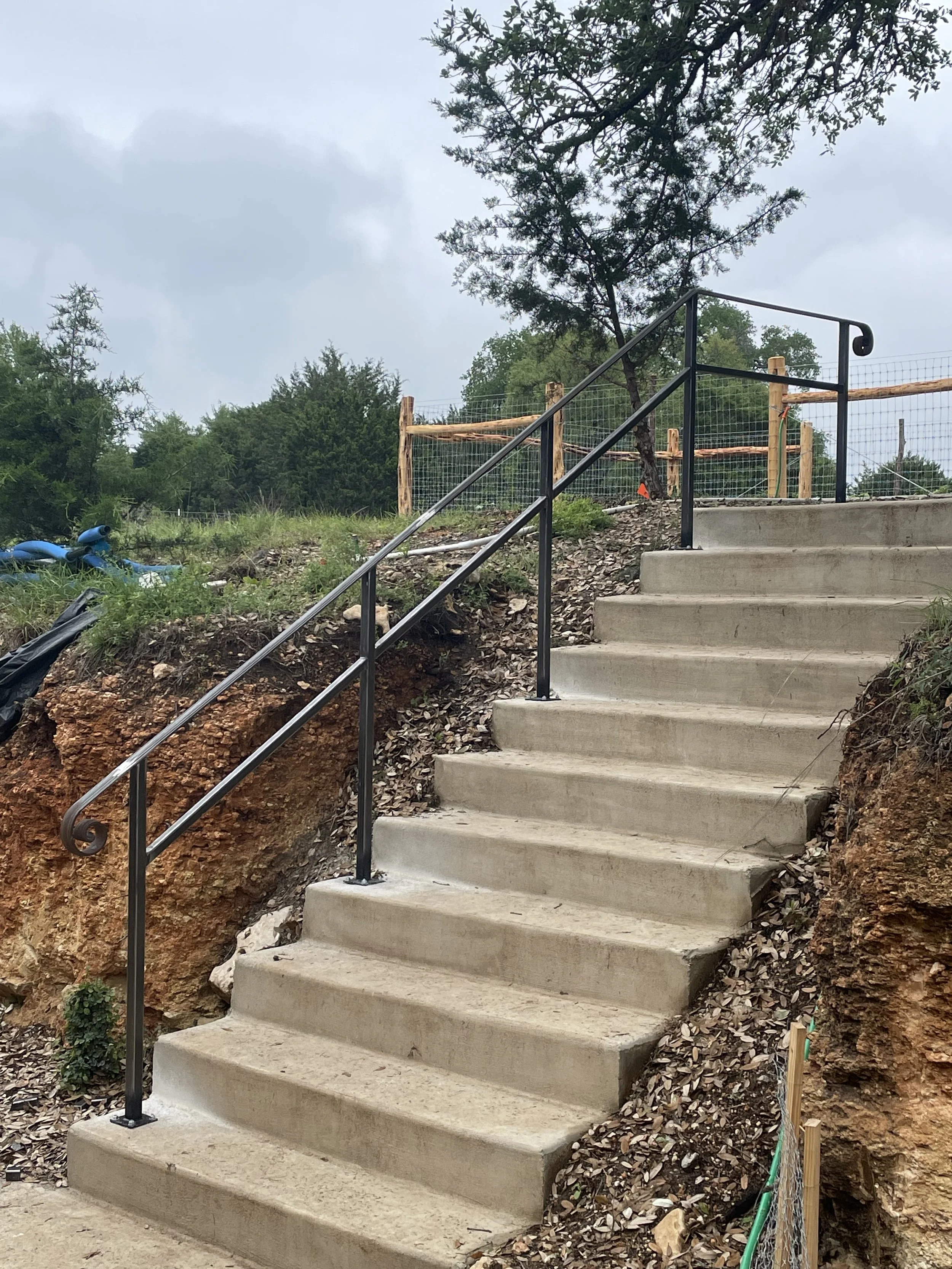 Concrete stairs with metal handrail beside an uneven dirt and rock slope, under a cloudy sky, with trees and construction fencing in the background.
