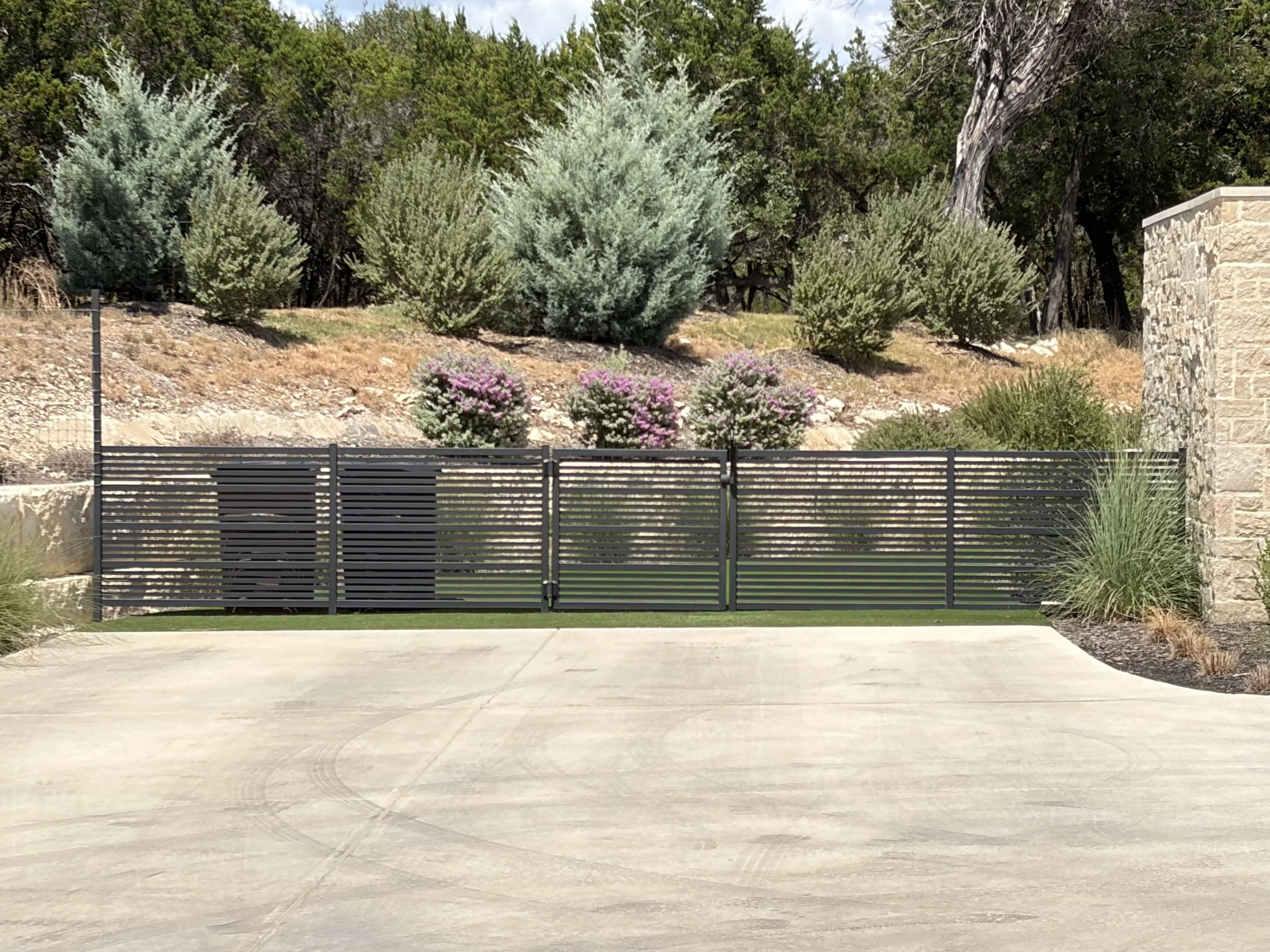 A landscaped area with a black metal screened enclosure with horizontal slats hiding trash can bins