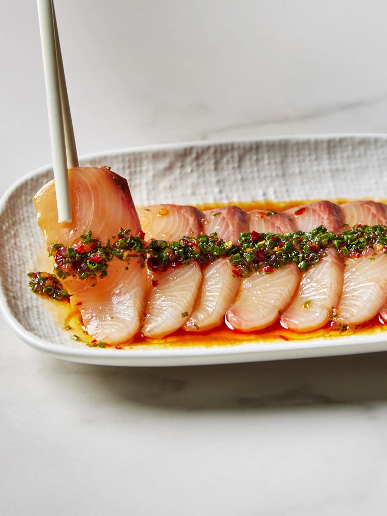Thinly sliced sashimi on a white plate, garnished with a spicy sauce and herbs, with chopsticks holding one piece.