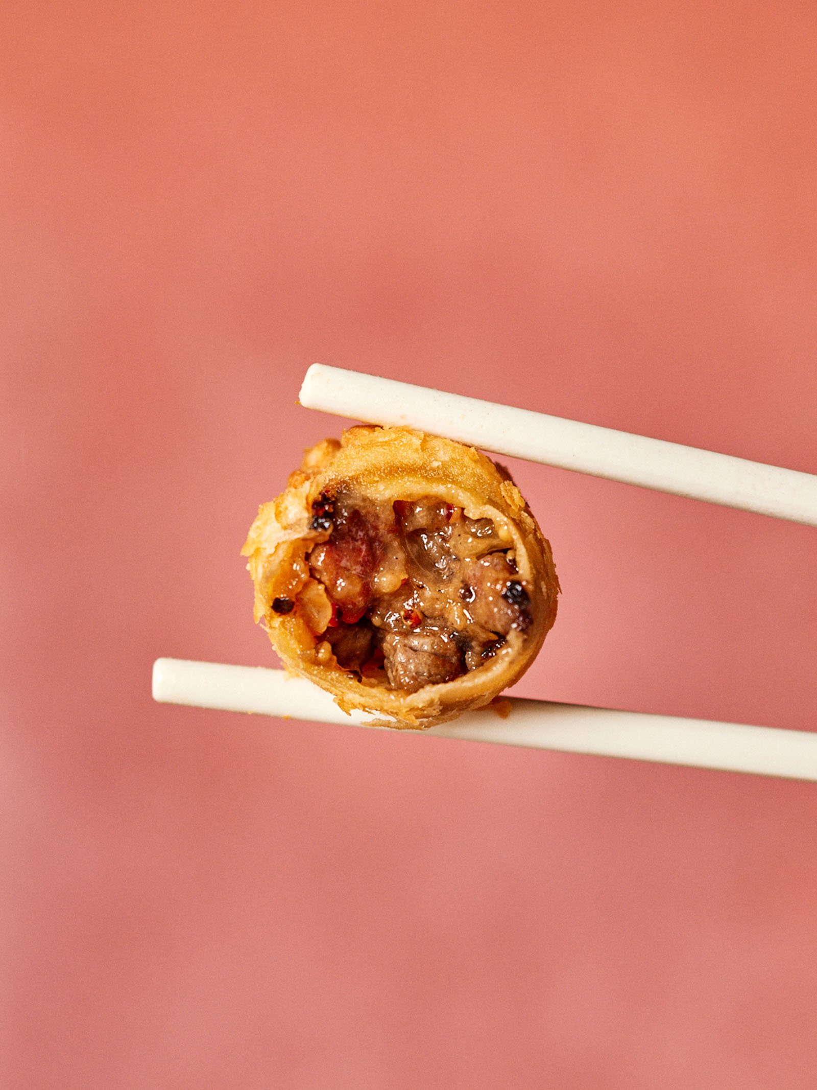 Chopsticks holding a cross-section of a crispy, filled food item against a pink background.