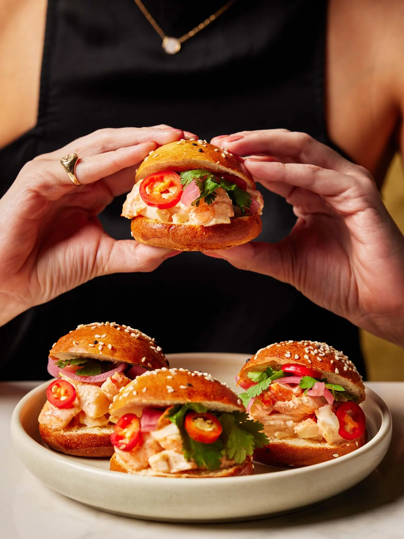 Person holding a Donna Chang Lobster Roll, with three similar sandwiches on a plate.