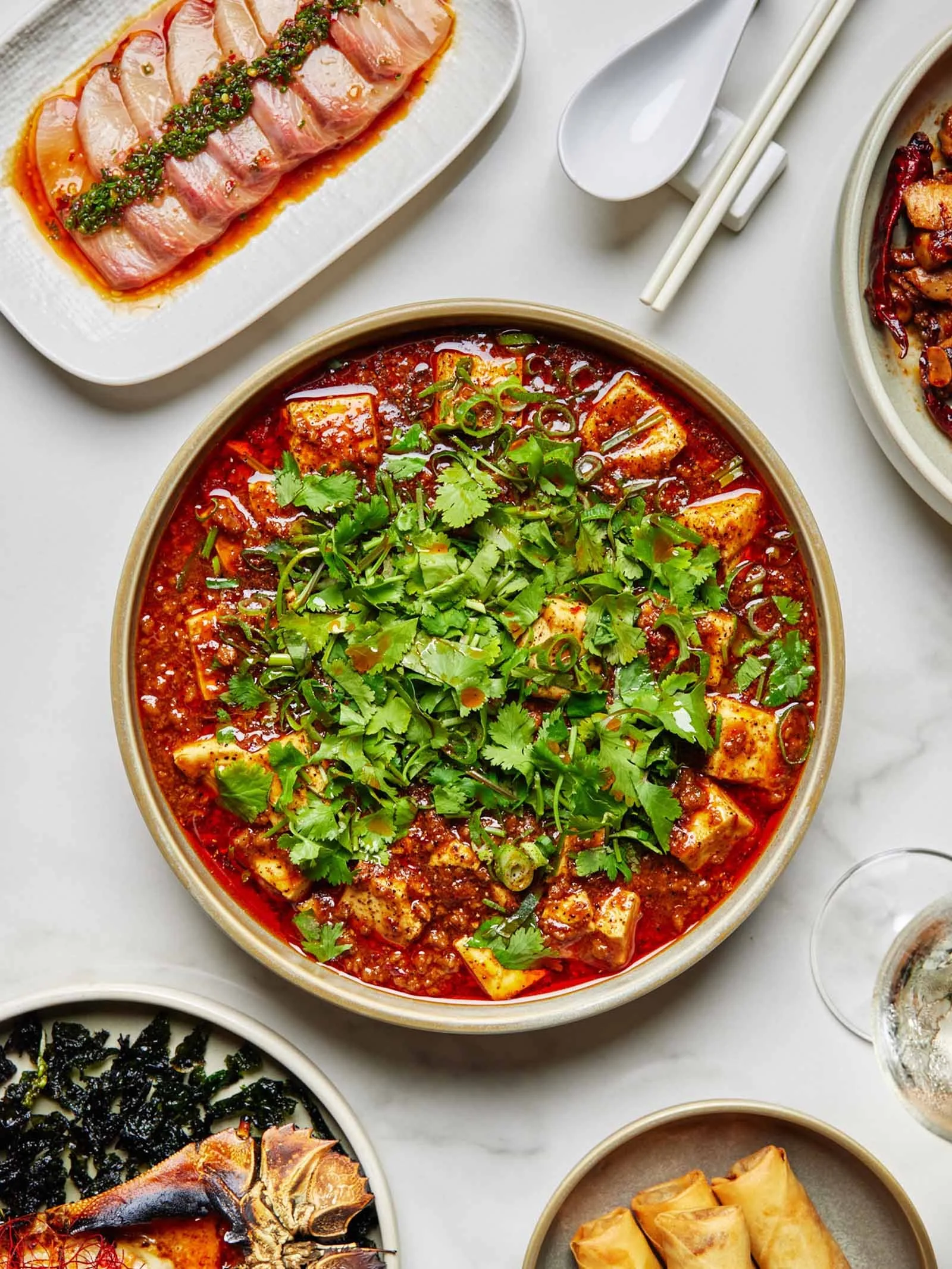 Asian dishes on a table including mapo tofu garnished with cilantro, a plate of sliced fish with sauce, a dish with seaweed and crab, and spring rolls. White chopsticks and a spoon are also visible.