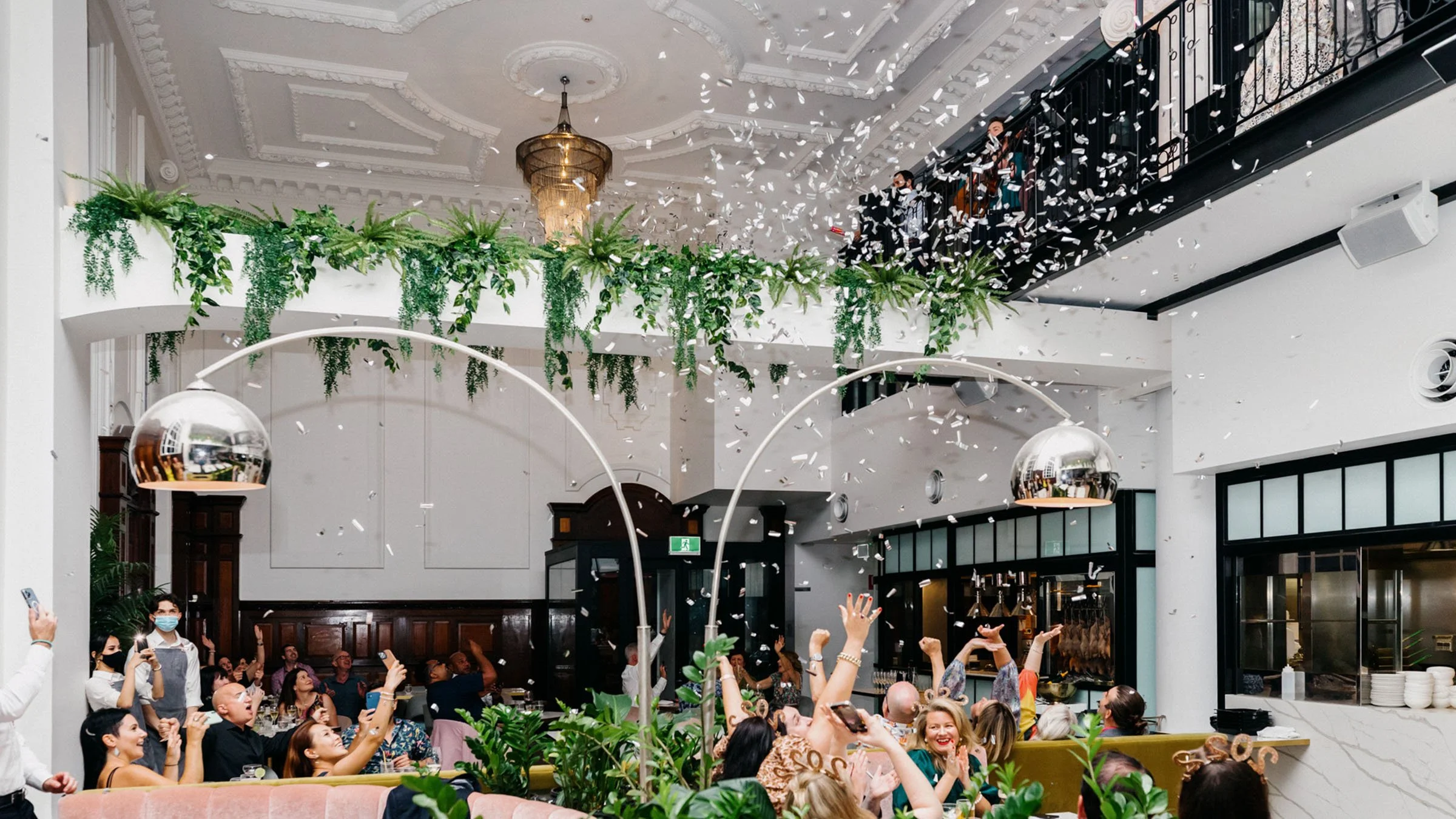 Indoor celebration with people cheering, confetti falling, and greenery decor in a stylish venue.