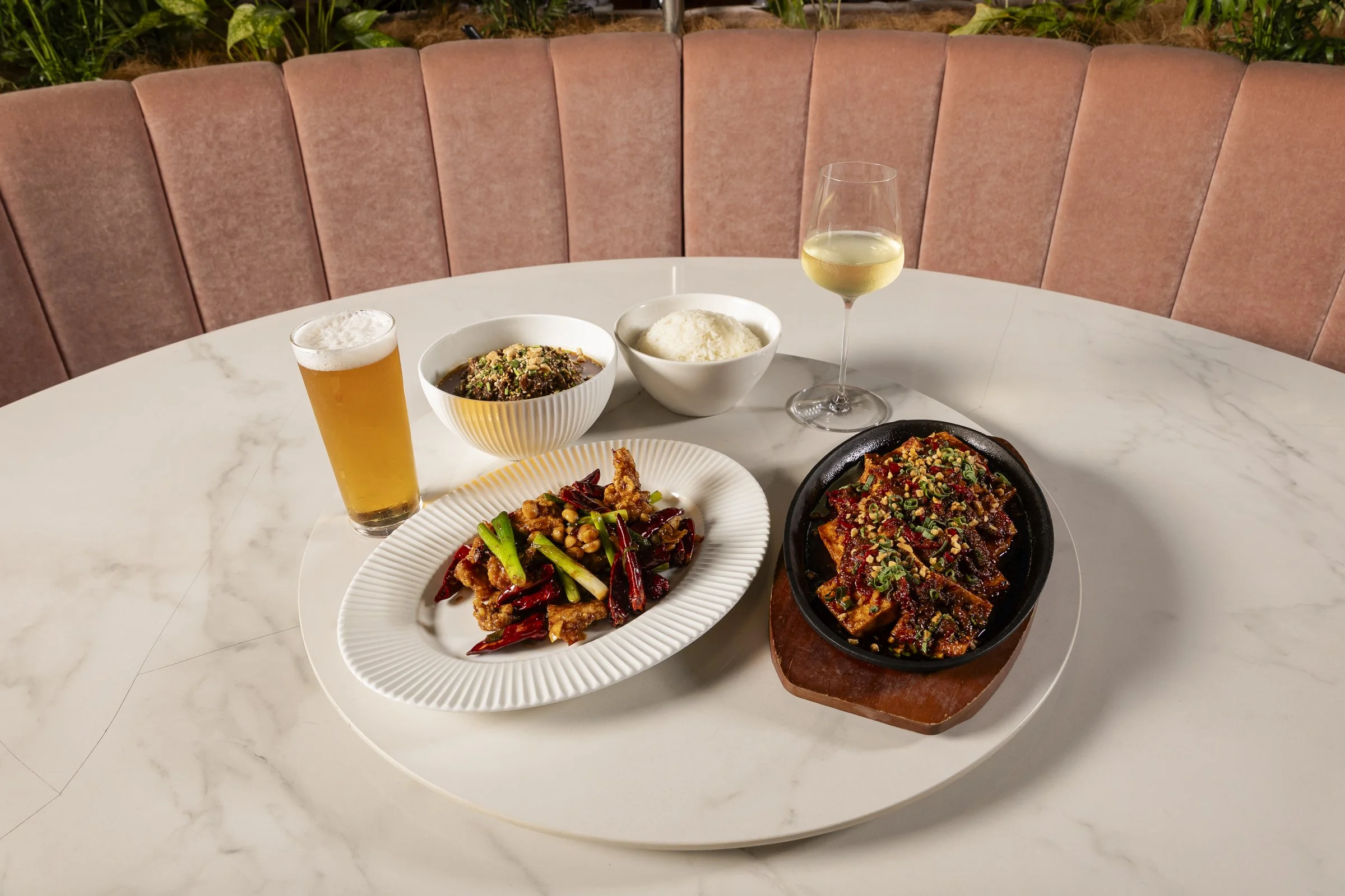 A round white marble table with various dishes including rice, stir-fried vegetables, and spicy tofu, along with a glass of white wine, a glass of beer, and a bowl of soup. Pink cushioned seating with tropical plants in the background.