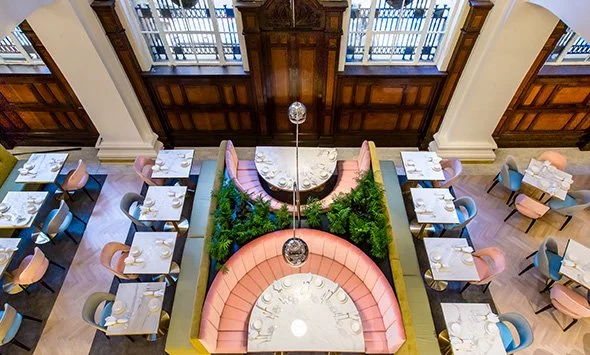 Aerial view of Donna Chang restaurant interior with circular booths, tables, and chairs in pastel colors. The booths have pink upholstery, and green plants are placed nearby. The wooden panel and large windows are in the background.