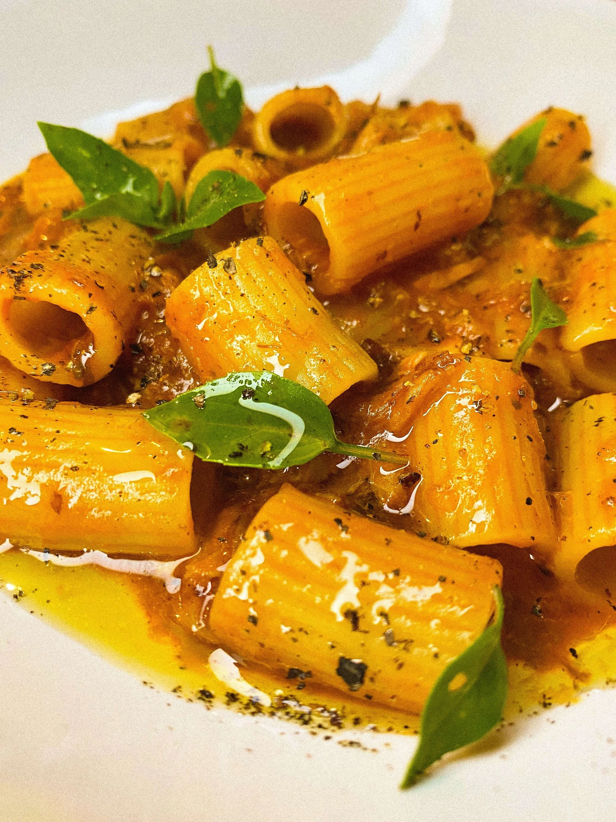 Close-up of cooked rigatoni pasta with herbs and spices, garnished with green leaves, on a white surface.