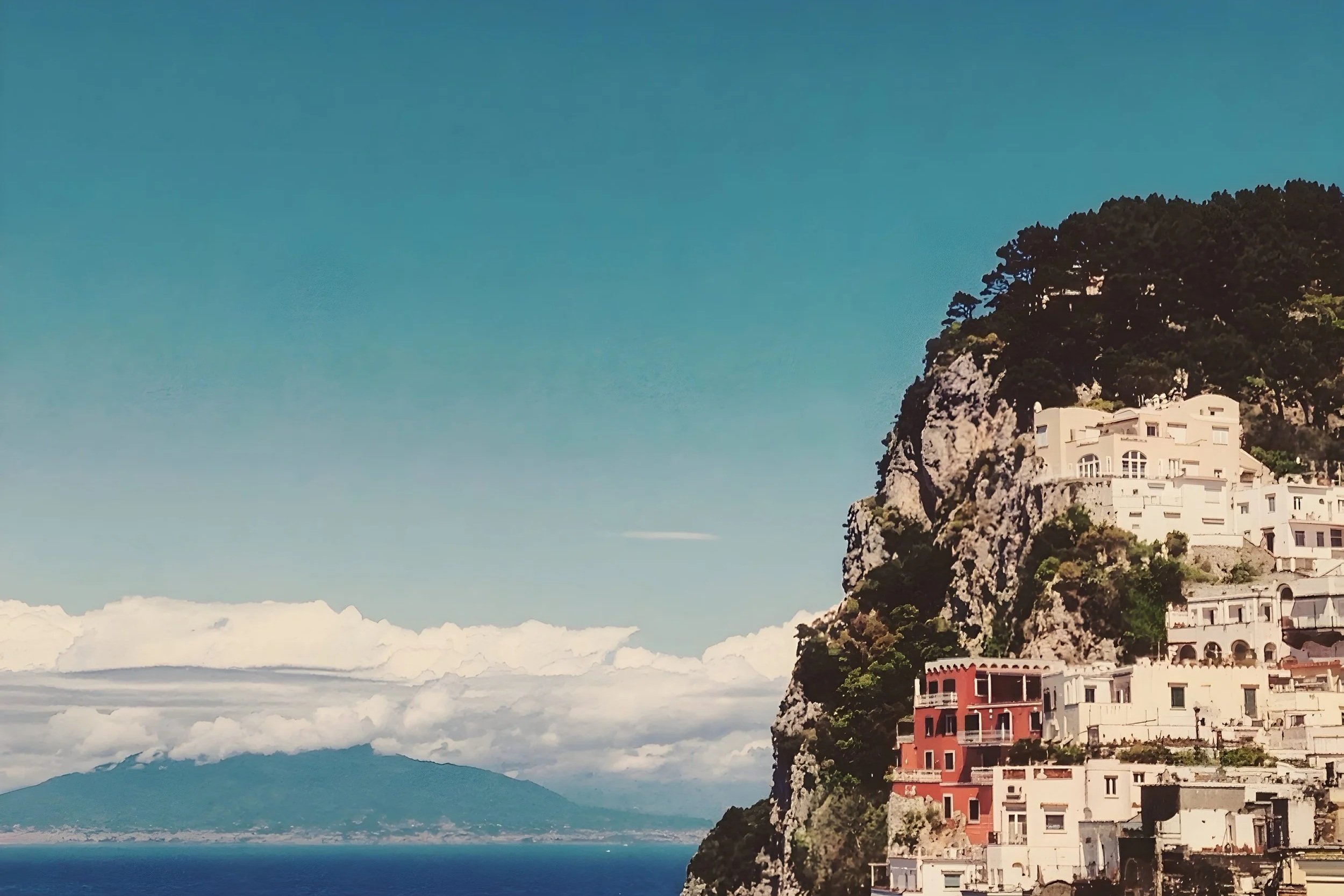 Coastal scene with a cliffside town of white and red buildings on a steep rocky hill, overlooking a body of water with distant mountains under a clear blue sky with white clouds.