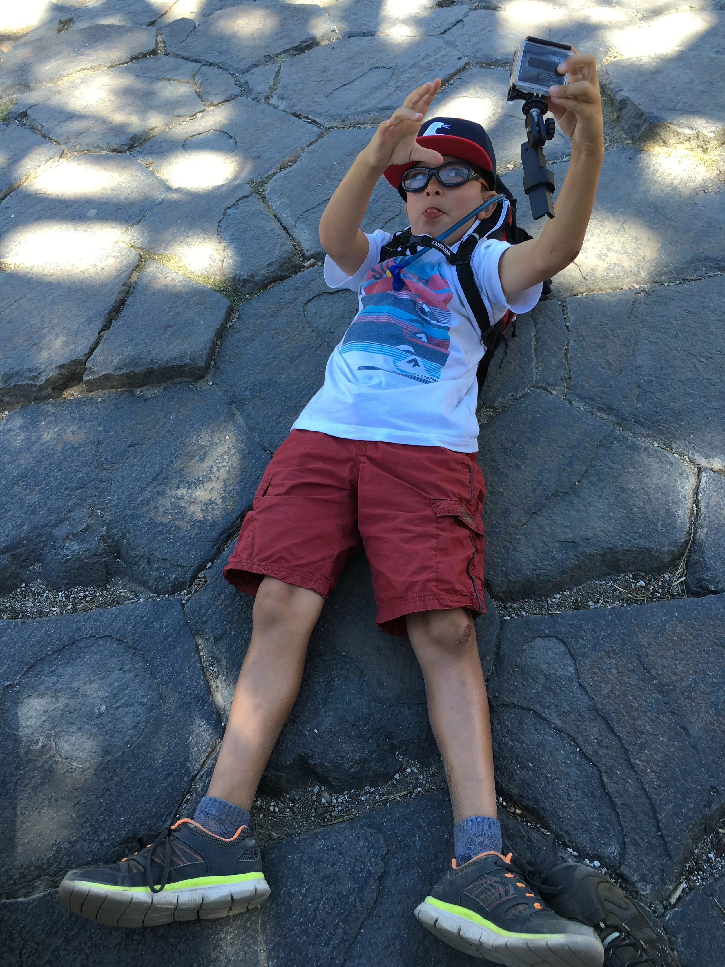 A young boy lying on his back on a stone pathway, taking a selfie with a GoPro camera, sticking out his tongue, wearing sunglasses, a white T-shirt with a colorful design, red shorts, and a red and navy cap.