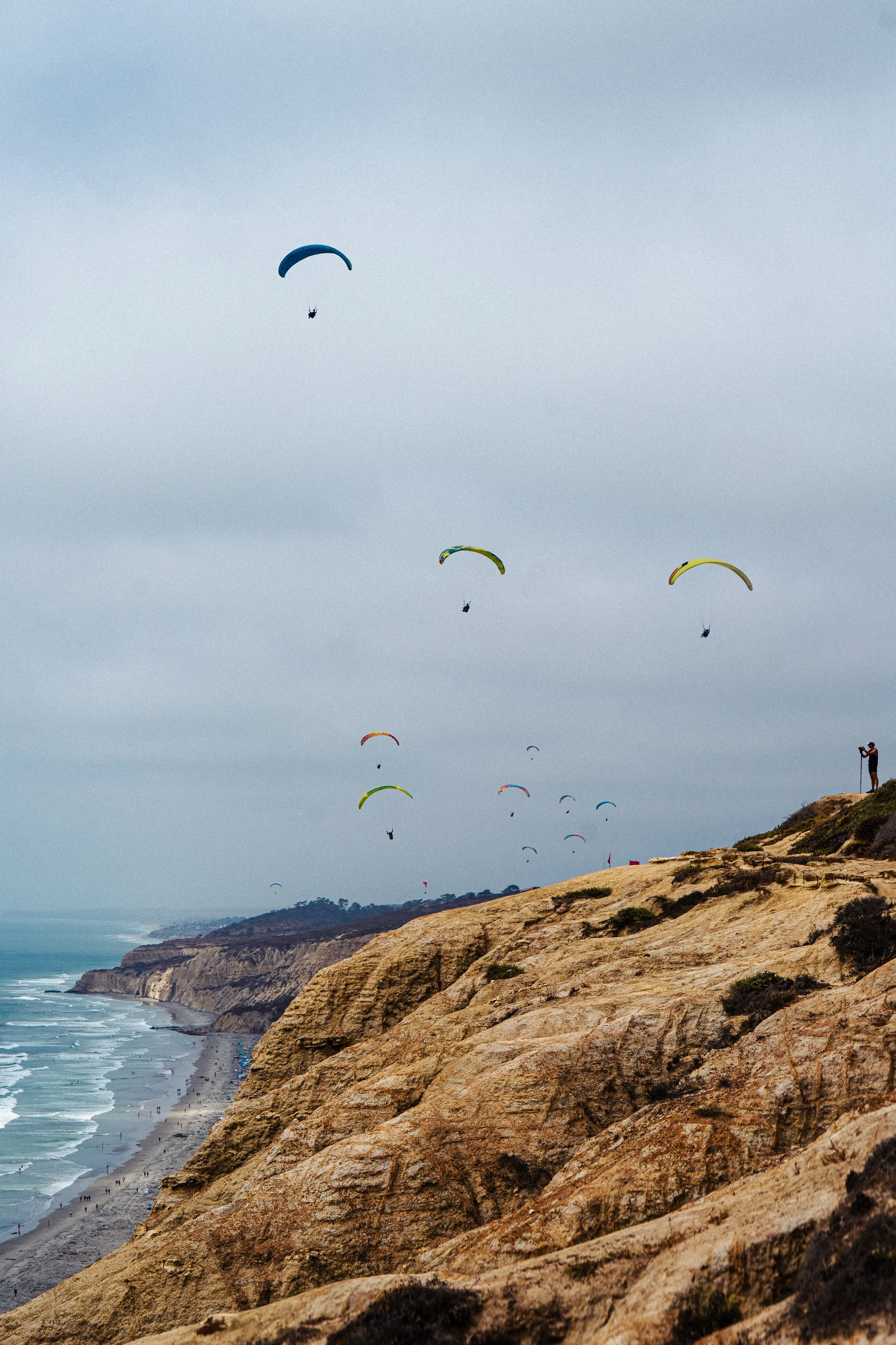 Multiple paragliders flying over a coastal cliff with a beach below, under a cloudy sky.