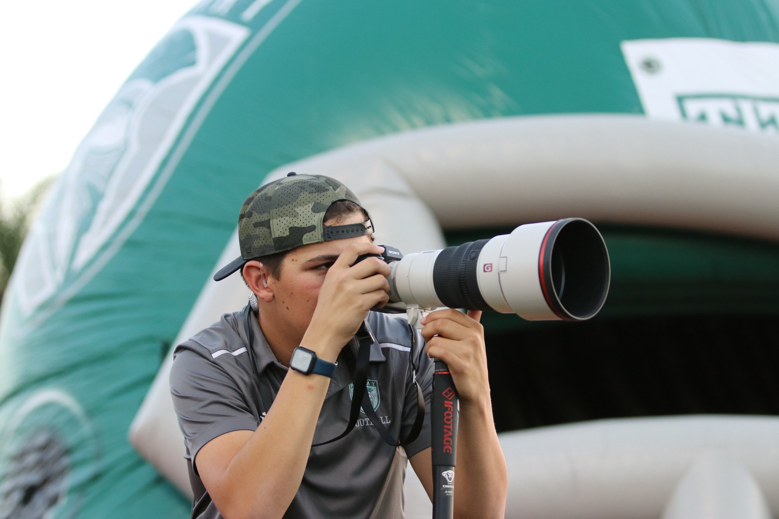 A young person in a gray polo shirt and a camouflage baseball cap is looking through the viewfinder of a large white camera with a long lens. They are standing outdoors under a green and white inflated structure, possibly an arch or tent.