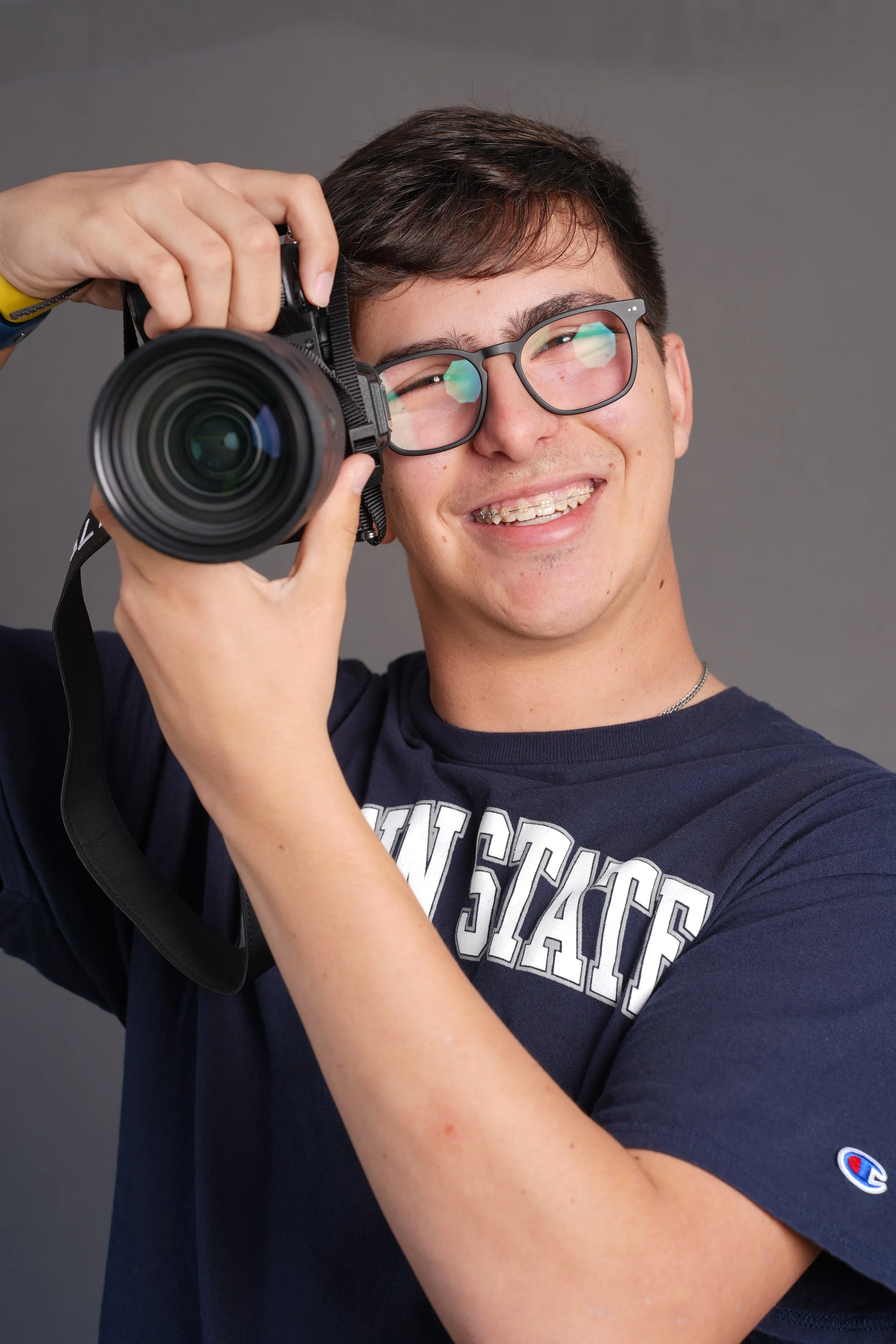 Young person with glasses holding a camera up to their face, smiling, wearing a dark T-shirt with white lettering, against a gray background.