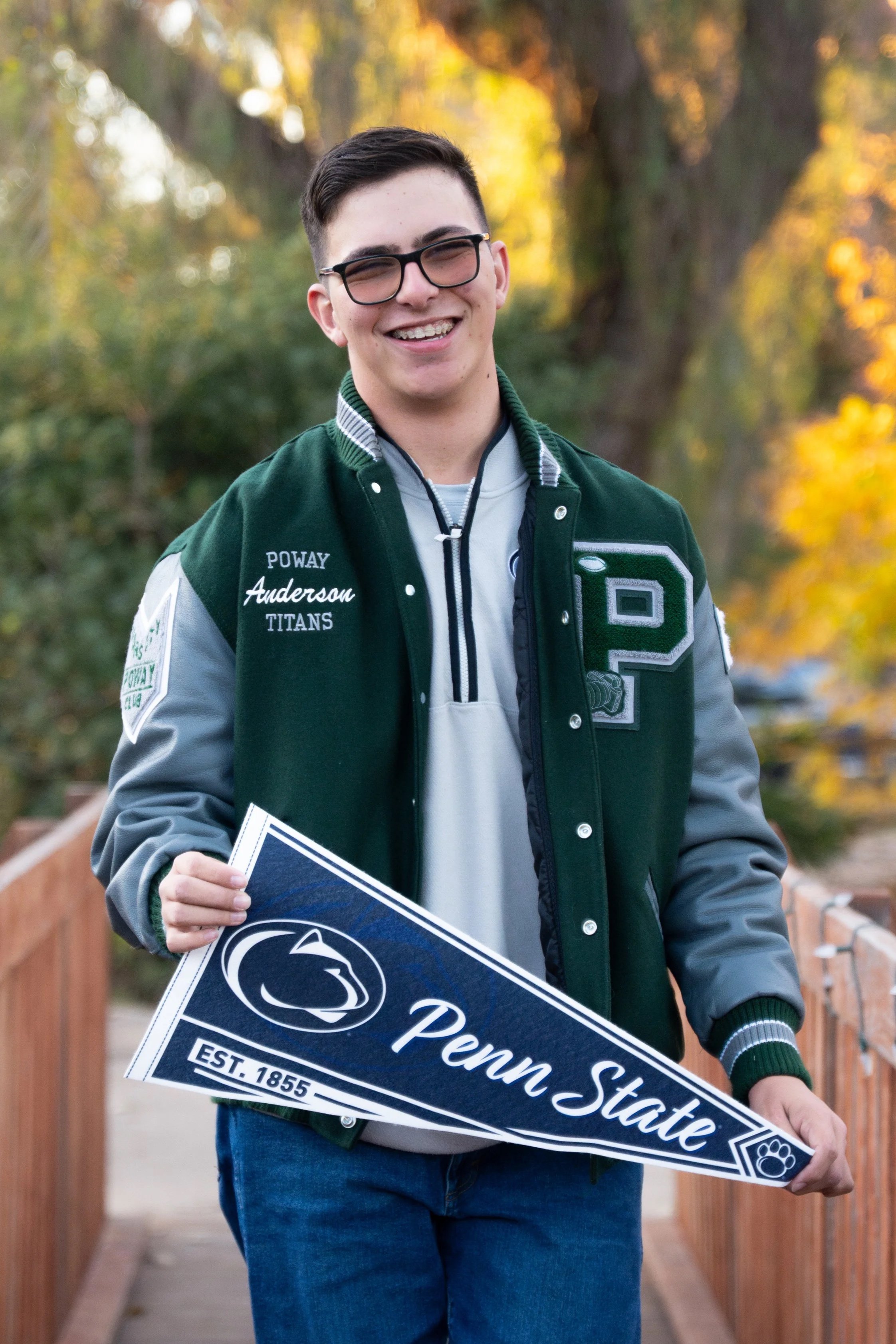 A young man walking outdoors on a wooden path, smiling, holding a Penn State football banner, wearing glasses and a green and gray jacket, with trees and warm fall colors in the background.