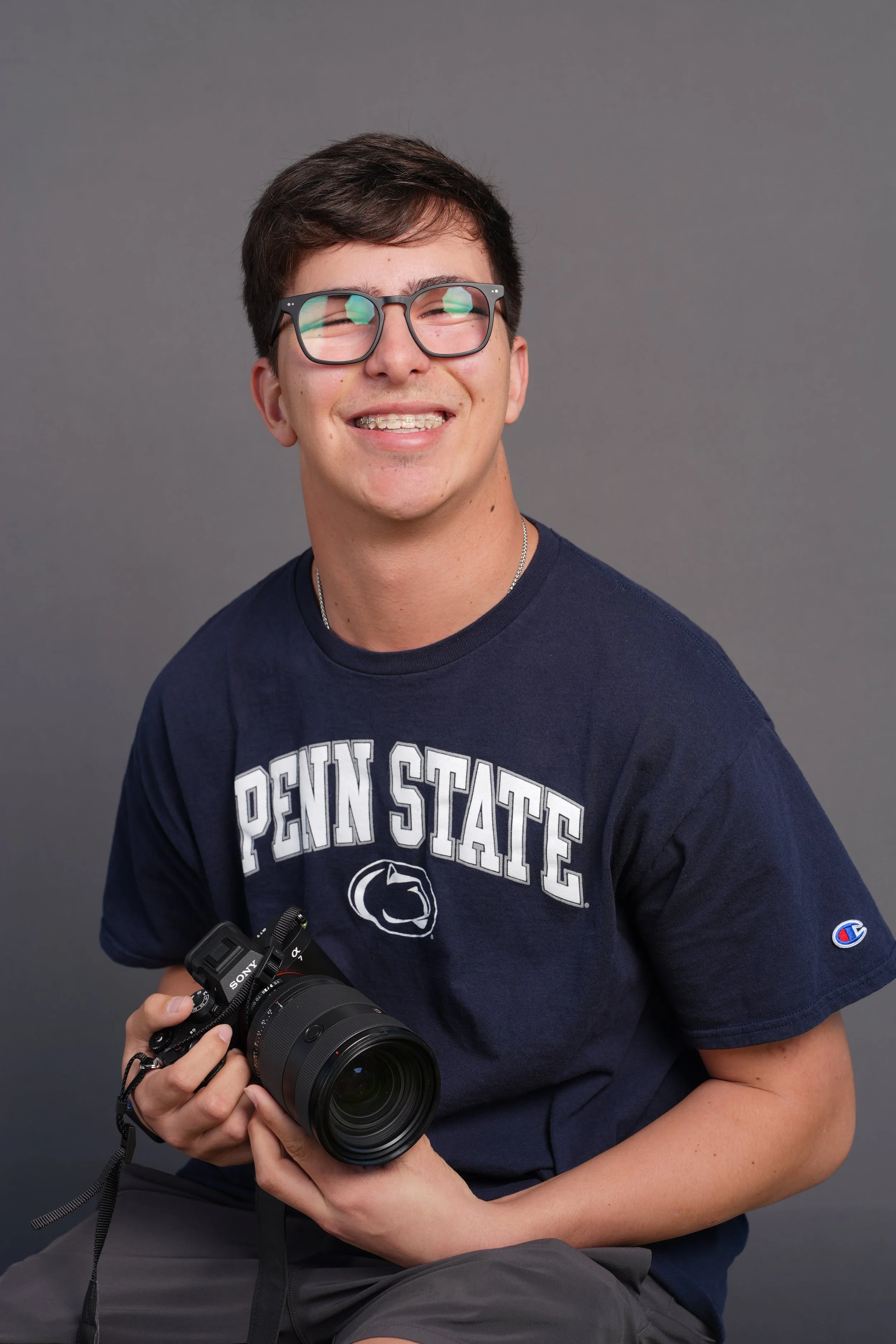 A young man with glasses, wearing a Penn State shirt, smiling and holding a camera against a plain gray background.