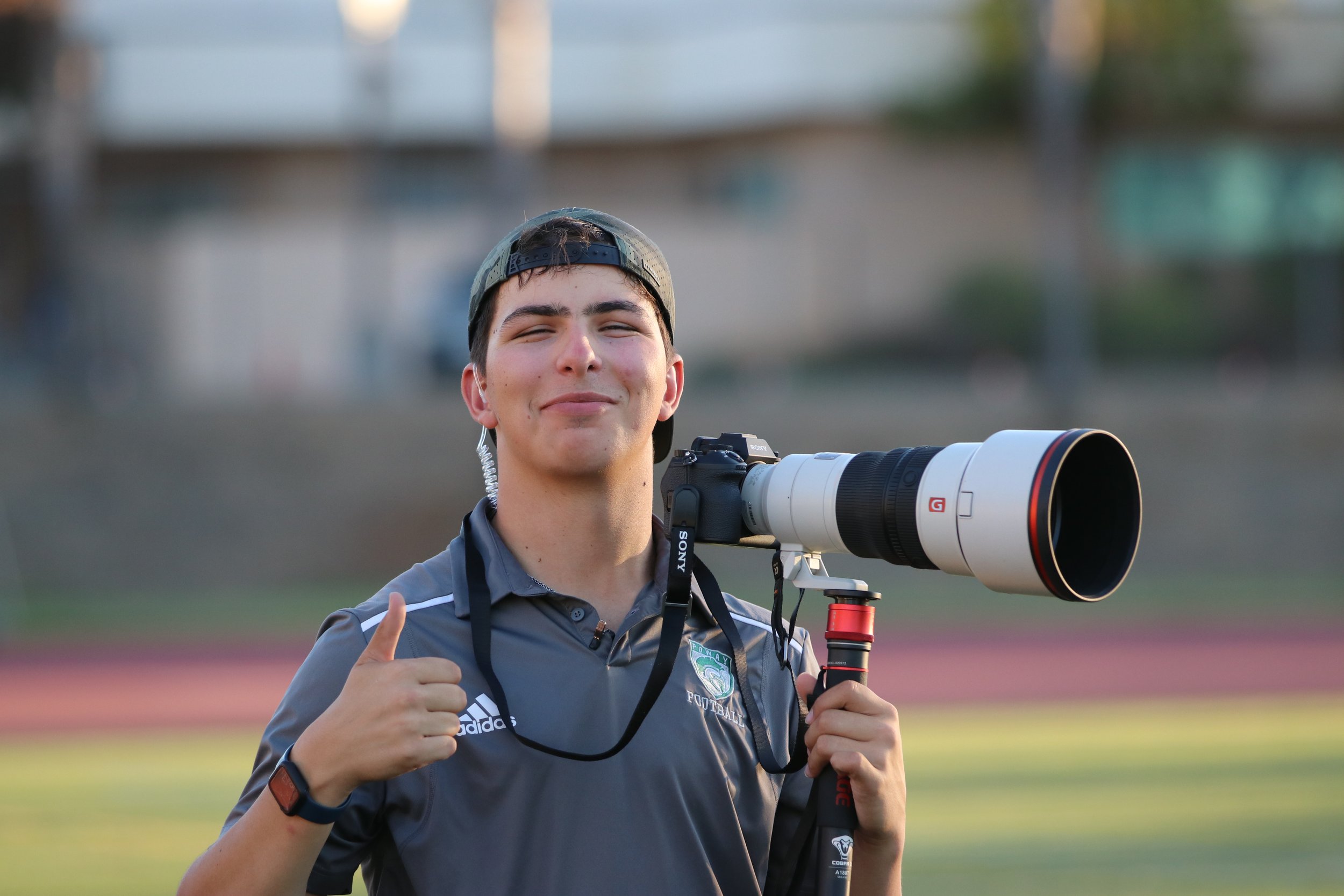 Young man smiling and giving a thumbs up while holding a professional camera with a large telephoto lens on a tripod outdoors.