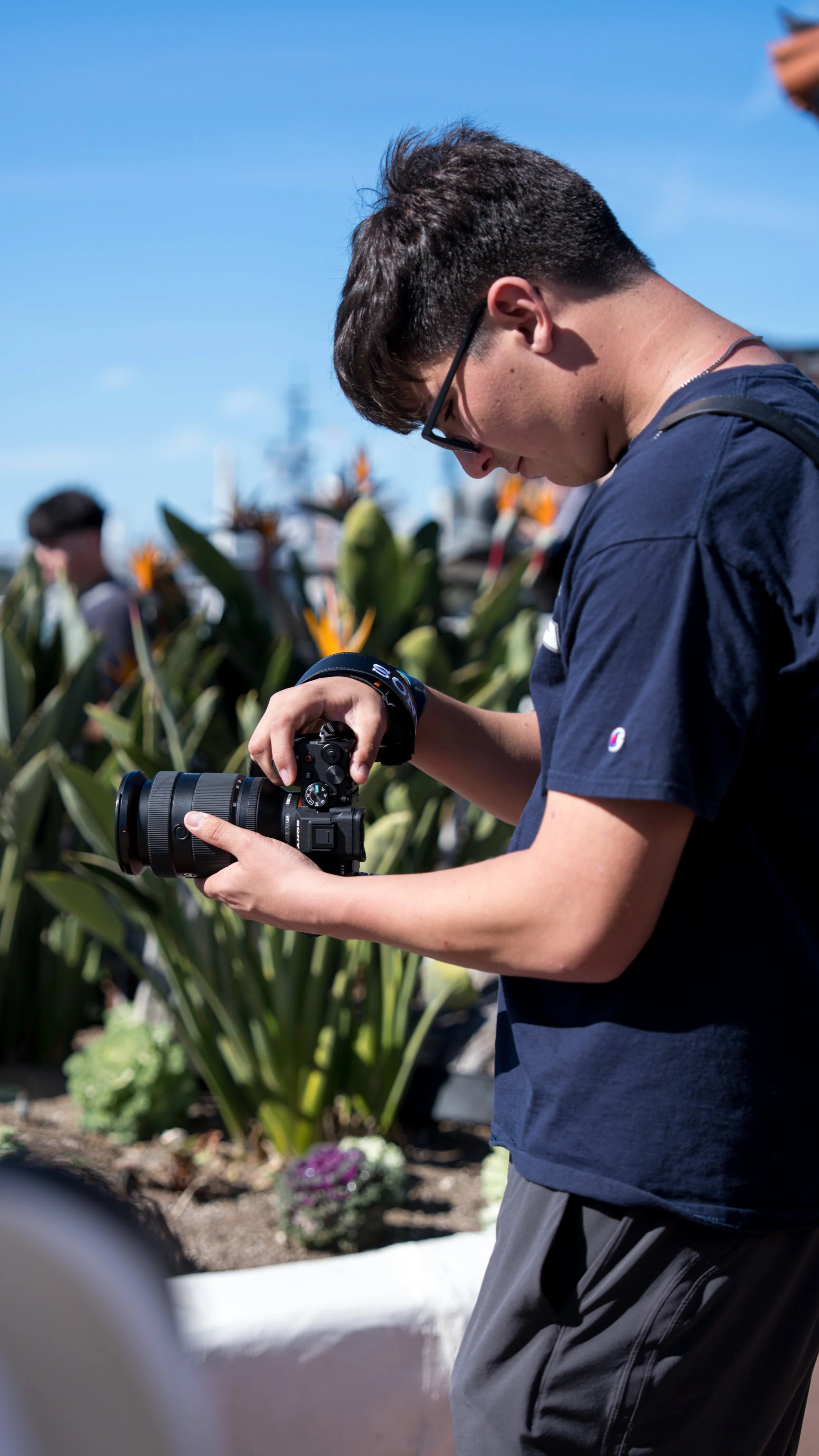 Young man with glasses using a camera outdoors on a sunny day, with green plants and a blue sky in the background.