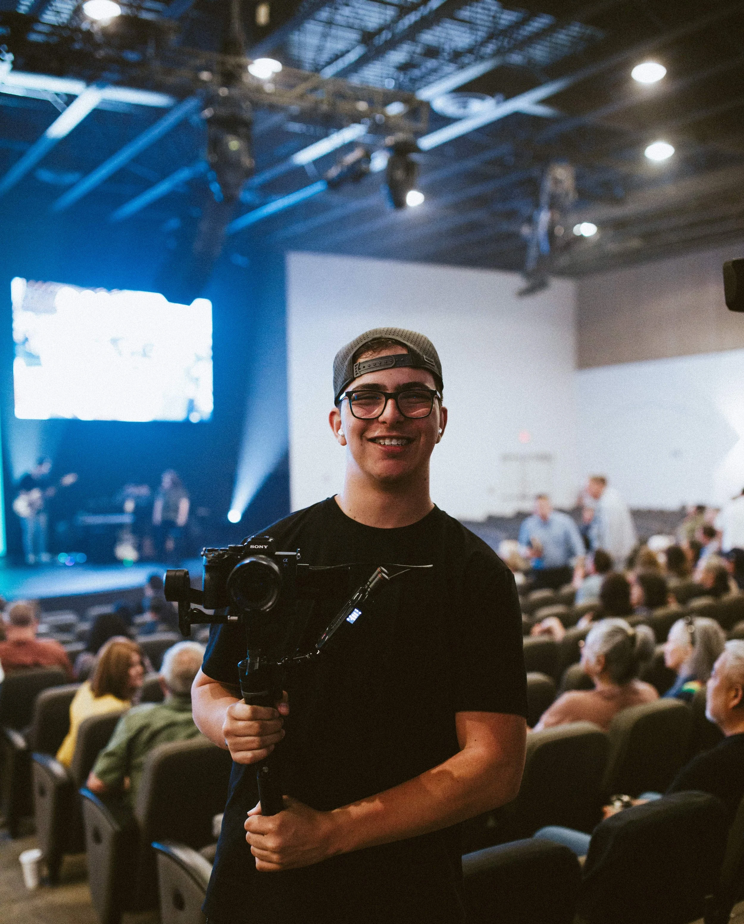 A young man wearing glasses and a backwards cap smiling, holding a camera on a stabilizer in a large indoor auditorium filled with seated people, stage, and overhead lighting.