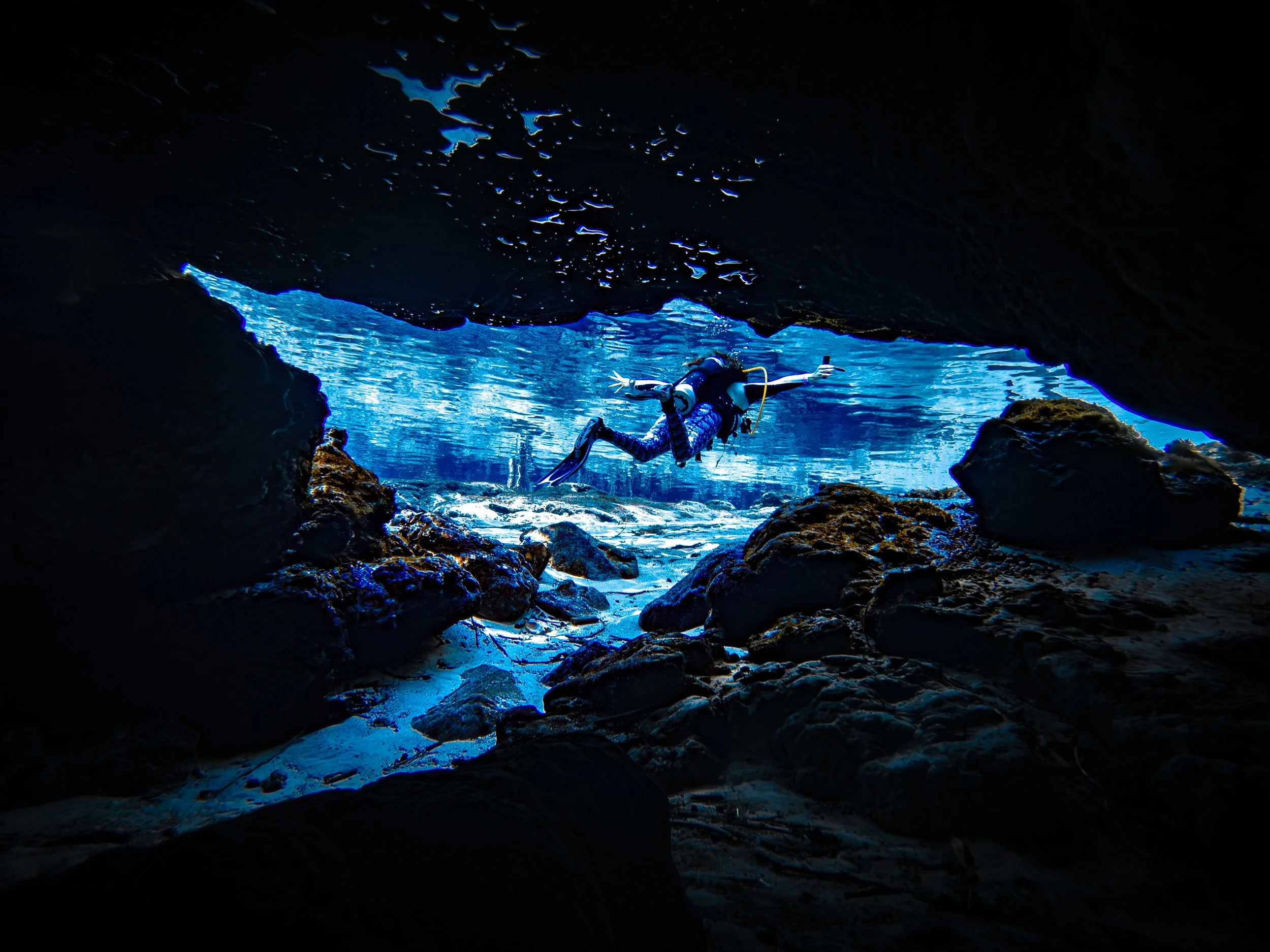 An open water diver in recrational scuba gear swims out of an underwater cavern at Ginnie Ballroom with dark rocky walls and blue water.