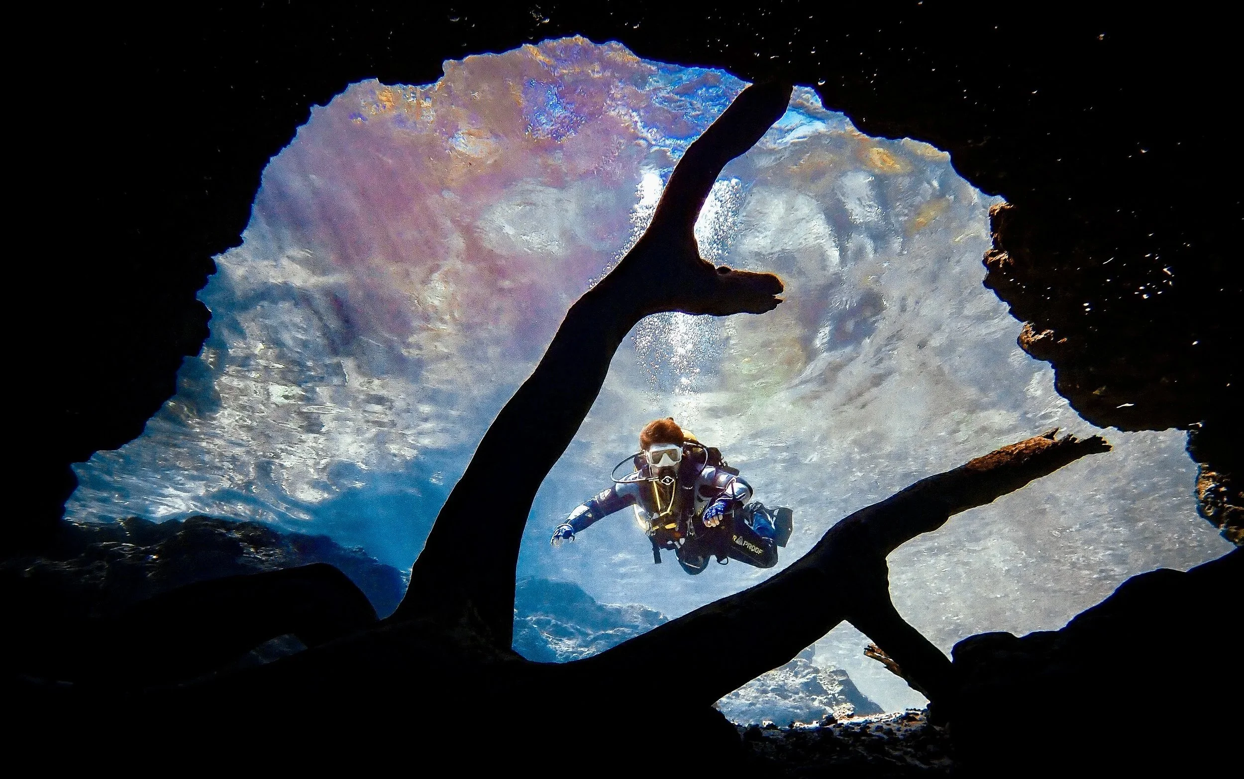 An underwater photograph of a scuba diver swimming between rocks, peering into the cavern zone at Little River, with the water surface visible above. The diver is wearing a wetsuit, mask, and fins, and is surrounded by bubbles and water.