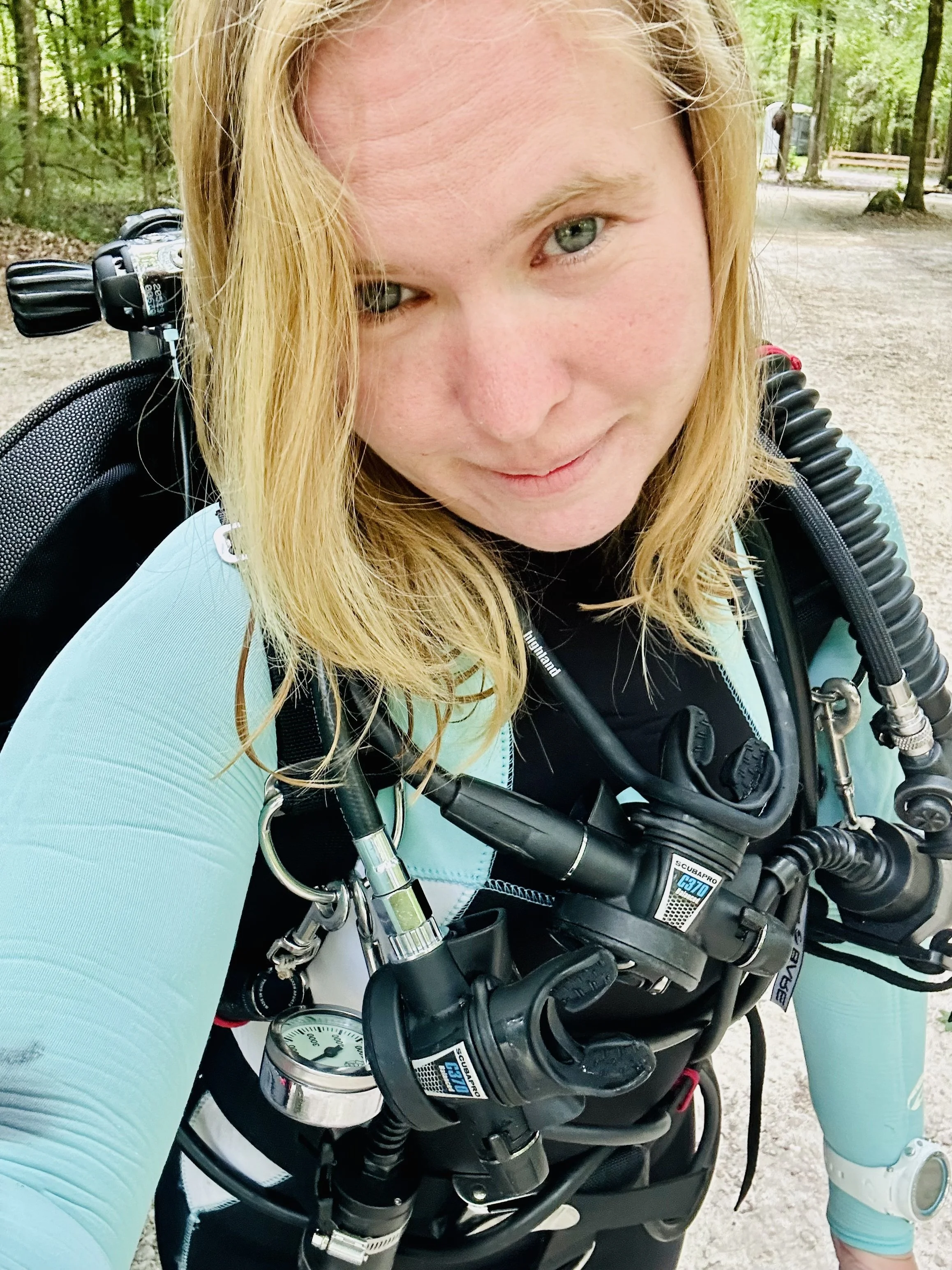 A woman with blonde hair wearing a teal wetsuit and backount doubles cave gear, standing outdoors in a wooded area.