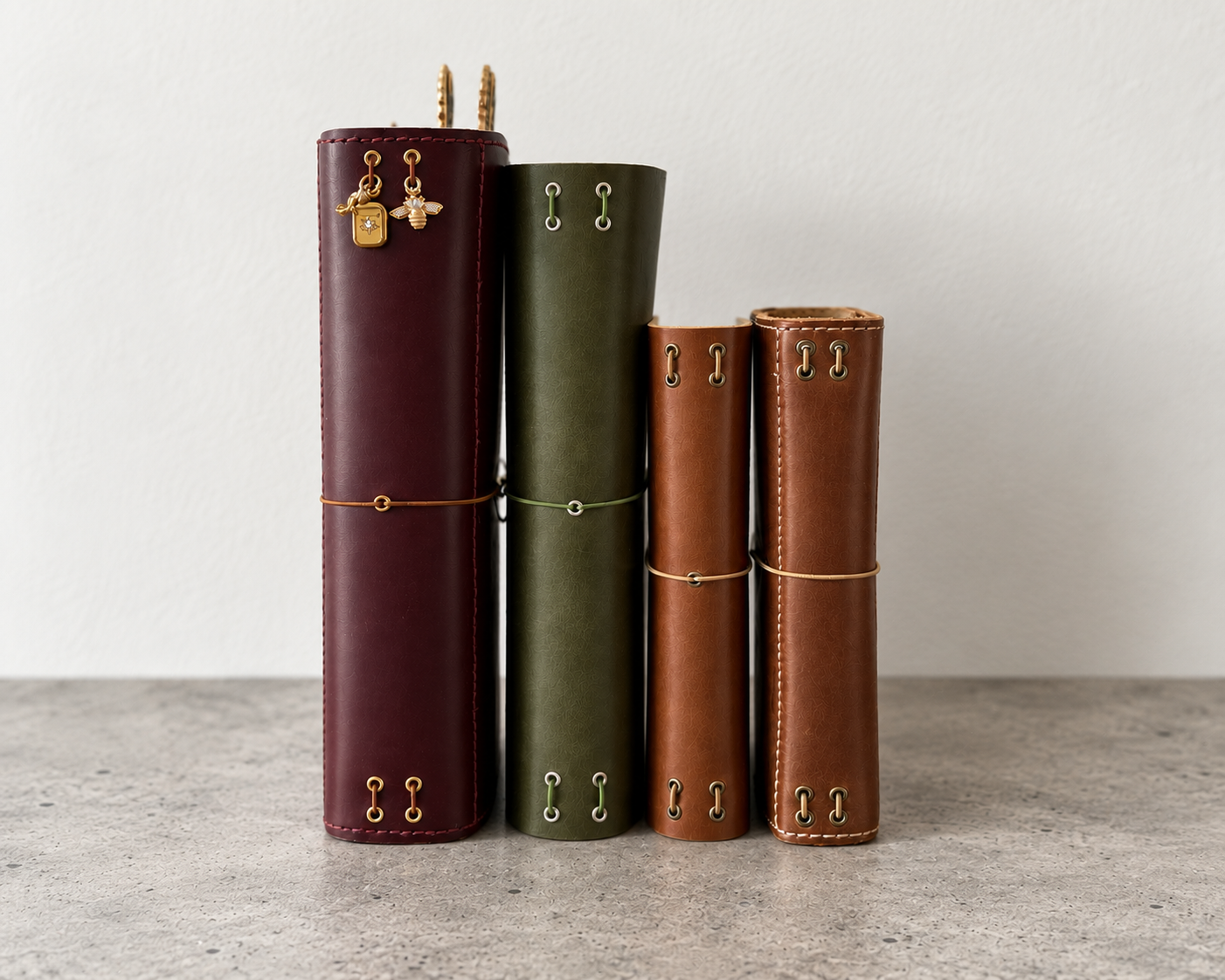 Four rolled-up leather journals in burgundy, green, and brown, standing vertically on a gray surface against a white wall, with pen charms attached to the top of some.
