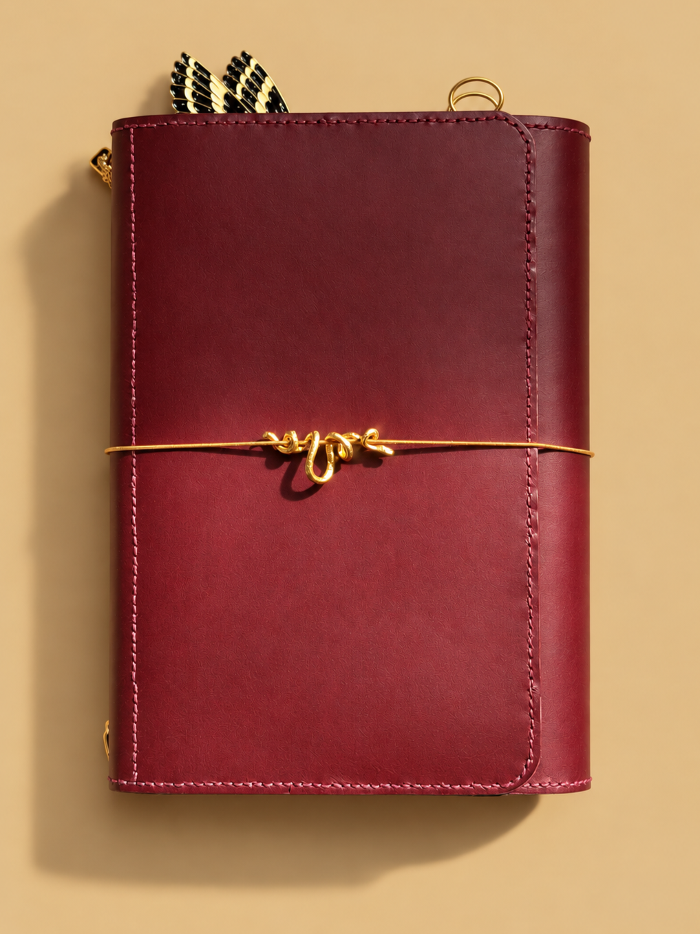 A closed maroon leather planner or notebook with a gold decorative elastic band wrapped around it, featuring a small snake tab embellishment in the center.