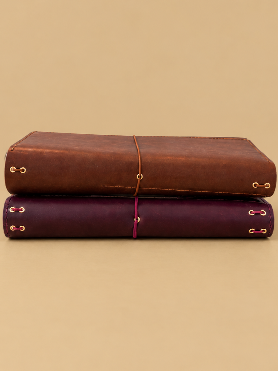 Two leather-bound books stacked horizontally, secured with colored strings, against a plain beige background.