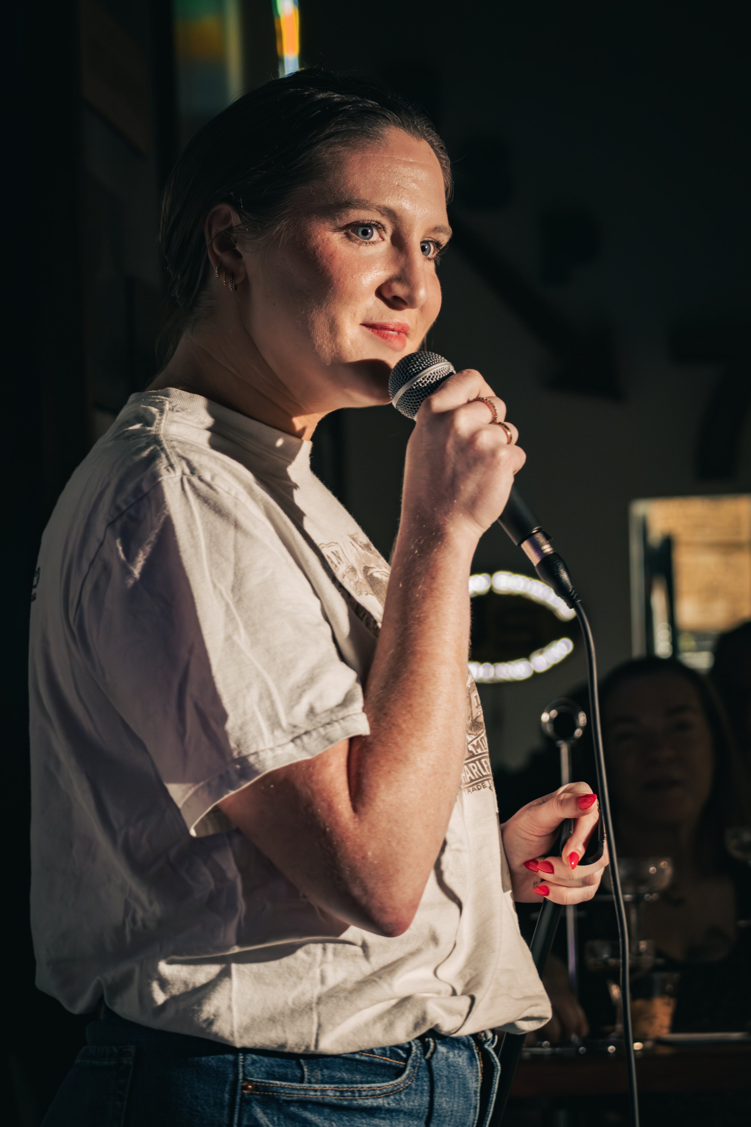 A woman with blue eyes and dark hair, wearing a white T-shirt, holding a microphone, appears to be singing or speaking on stage in a dimly lit setting.