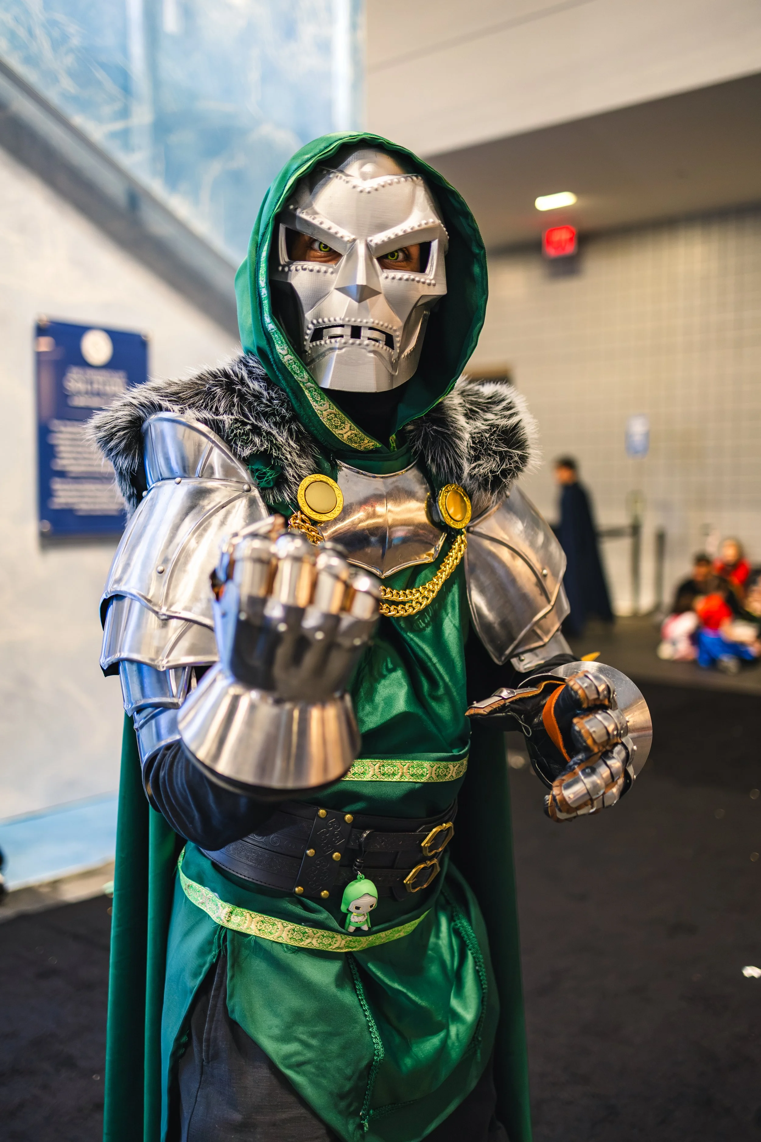 Person dressed in a detailed, metallic skull mask, green cloak, and armor, standing in an indoor setting with people in the background.