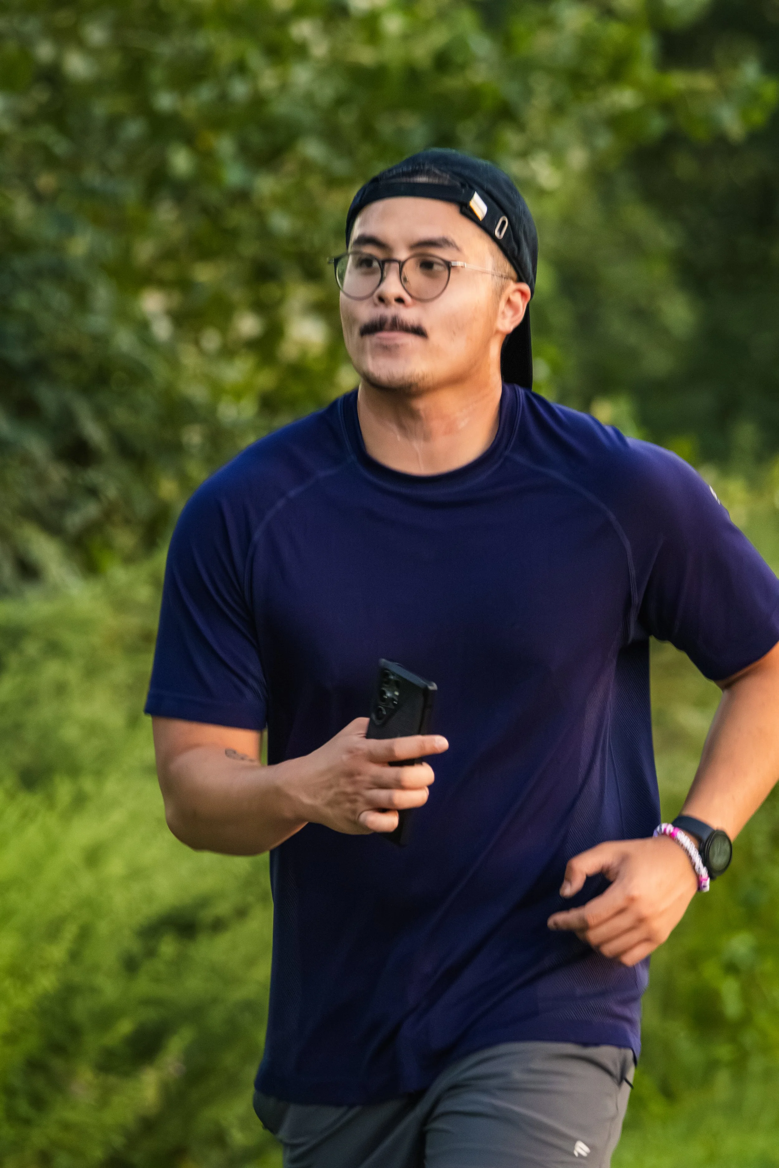 A man wearing glasses, a black cap, a navy blue athletic shirt, and gray shorts is running outdoors on a green grassy trail with trees behind him, holding a smartphone in his right hand.