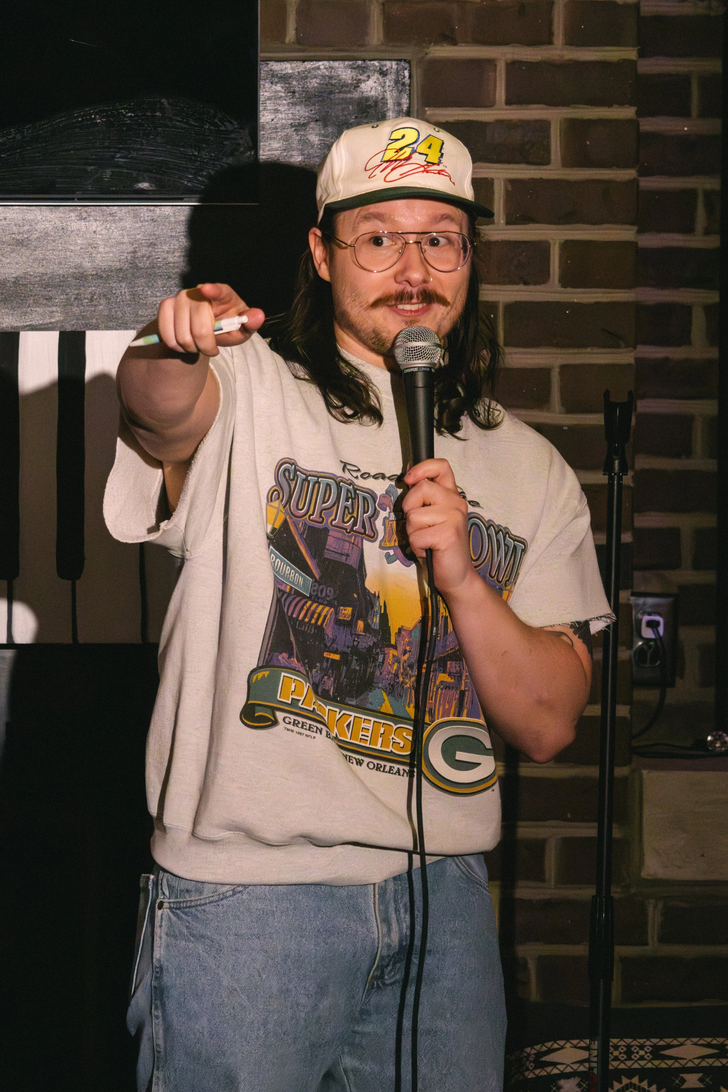 A man with long brown hair, glasses, and a moustache is speaking into a microphone. He's wearing a white cap with the number 24, a white t-shirt with a graphic that says 'Super Bowl', and blue jeans. The background shows a brick wall.
