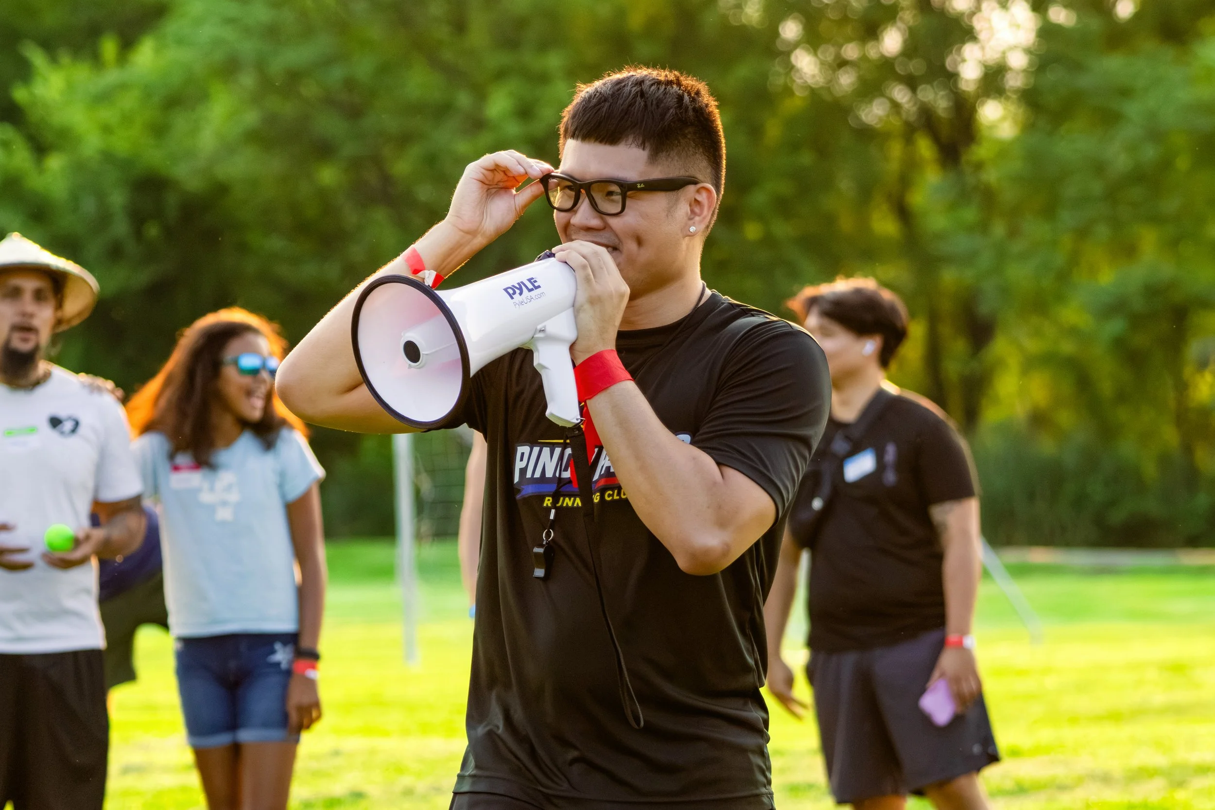 Man wearing black shirt, glasses, and a red wristband, holding a white megaphone with the brand DYLE, speaking to a group outdoors during daytime in a park with green trees, with other people in the background.