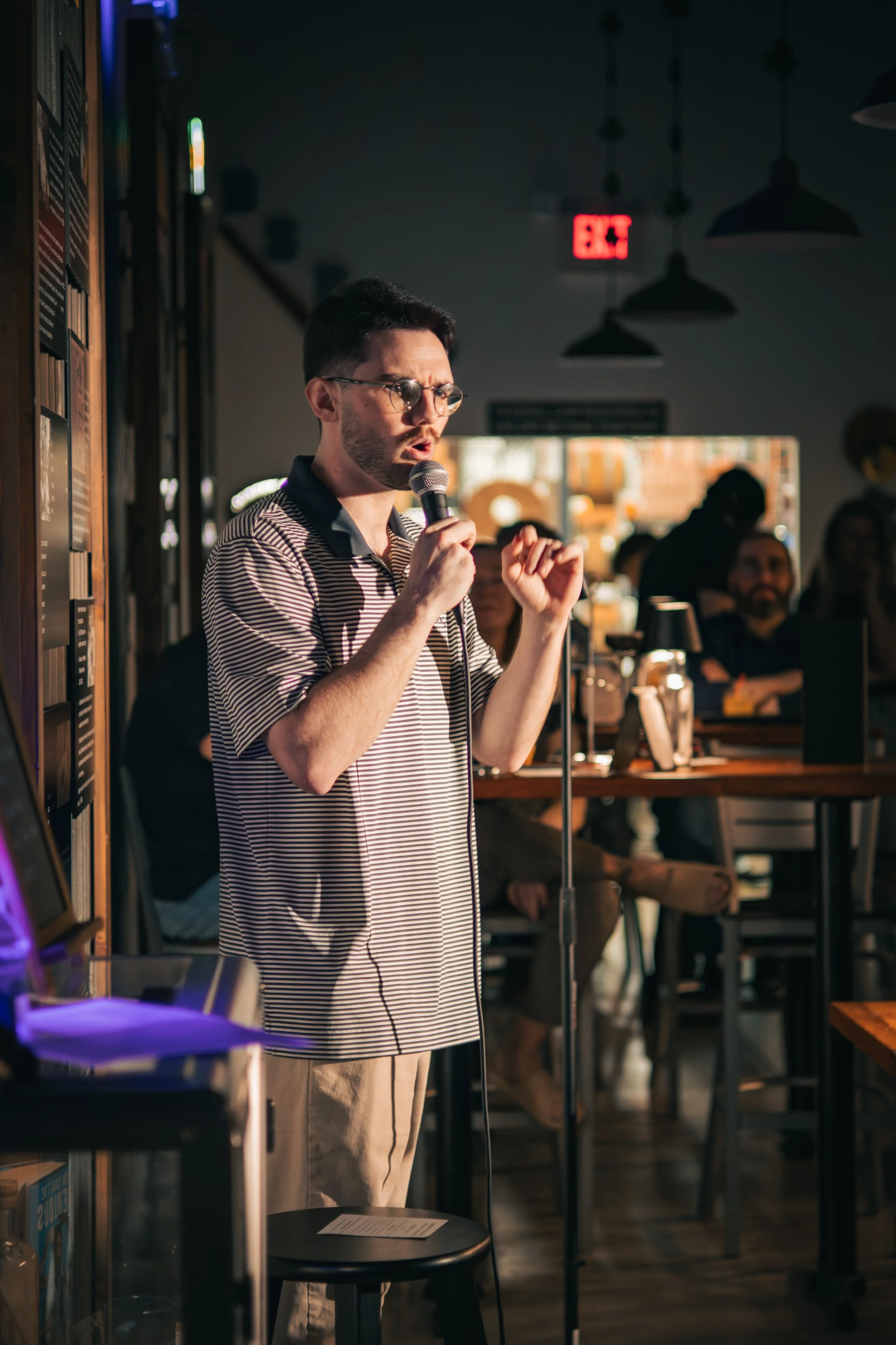 A young man with glasses singing into a microphone inside a cozy, dimly lit cafe. He is wearing a striped polo and beige pants, with people sitting at tables in the background.