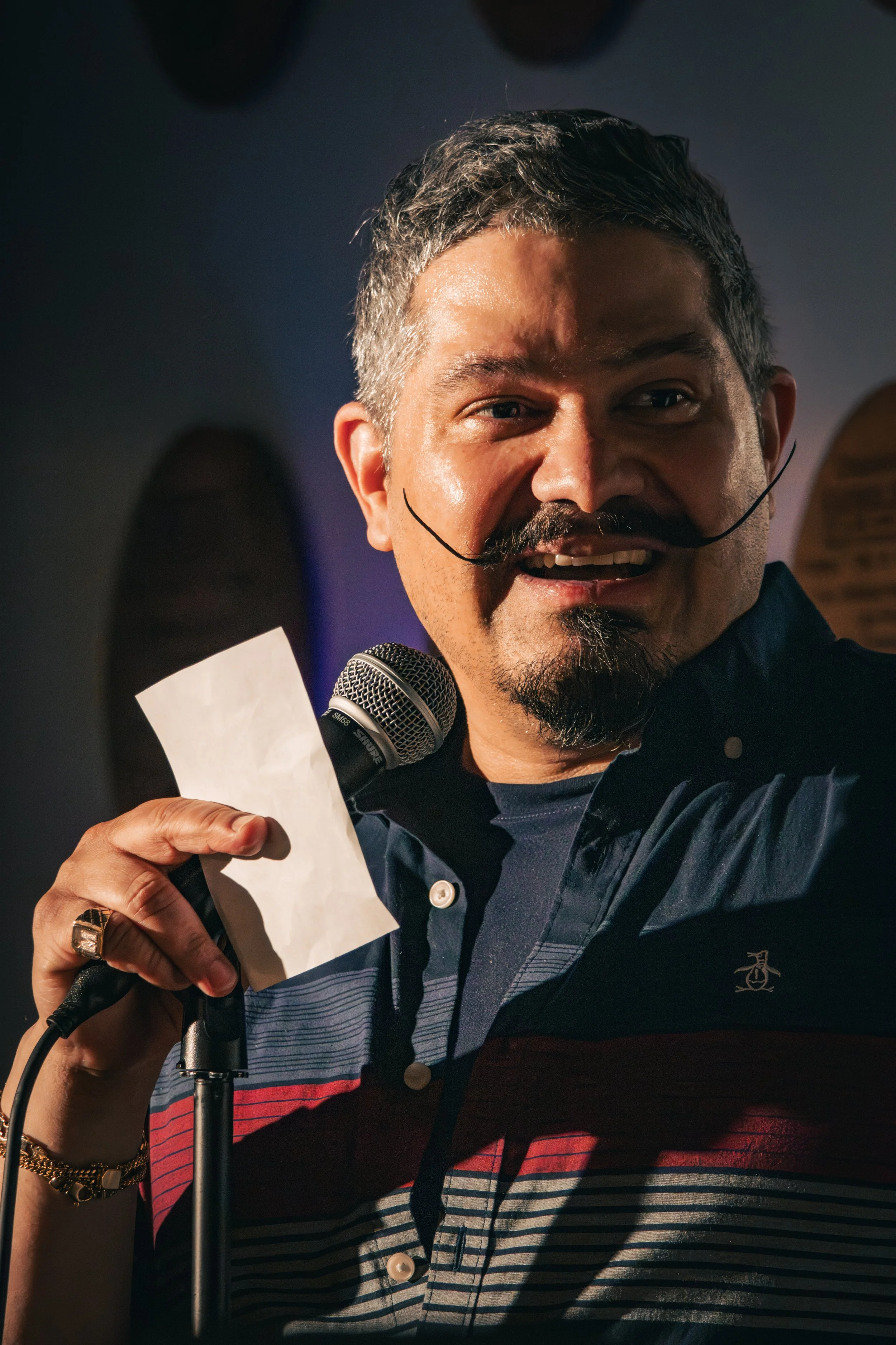 A man with short, dark hair and a mustache is speaking into a microphone while holding a piece of paper. He is wearing a striped shirt with a dark collar and appears to be indoors with a dark background.
